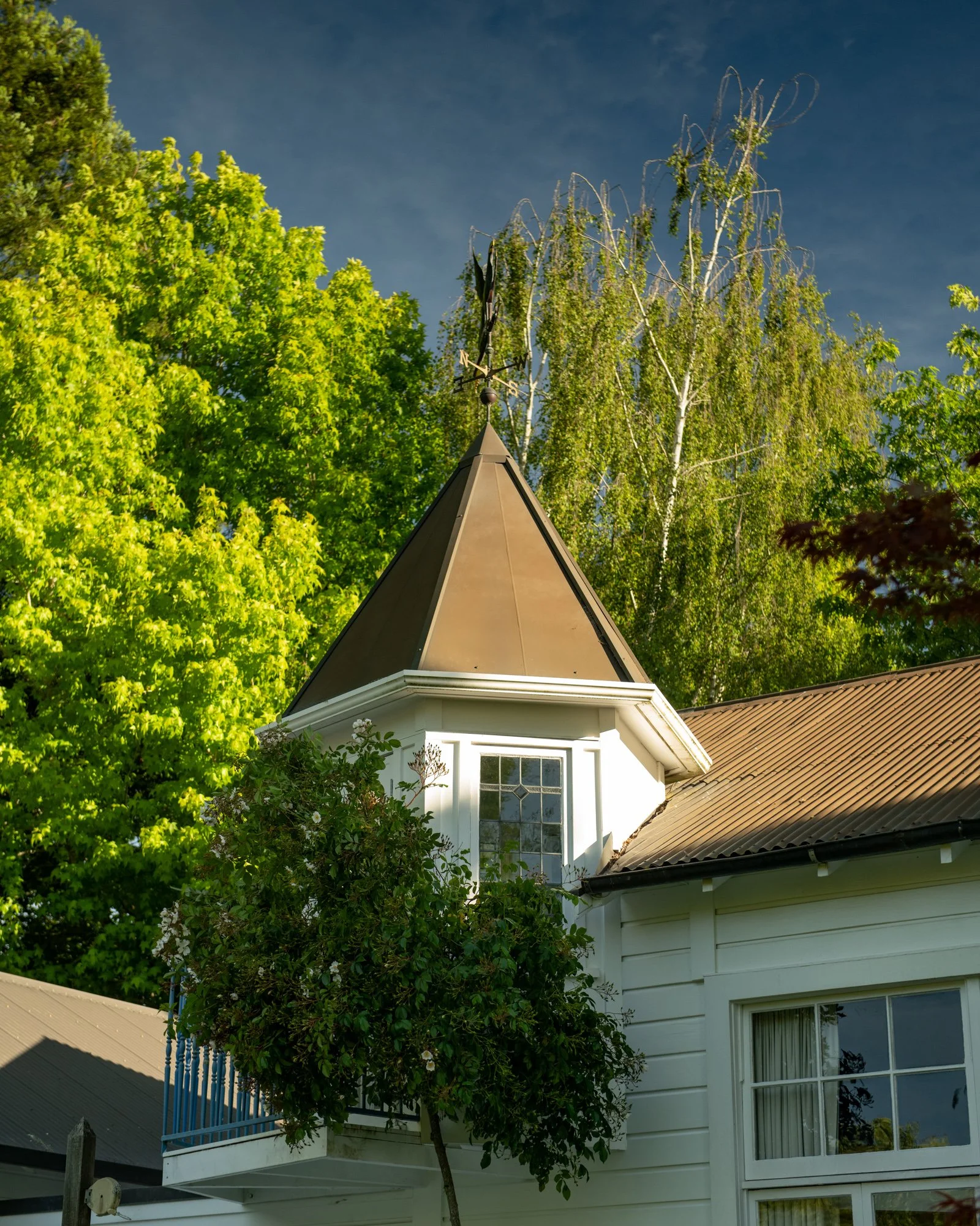A white house with a steep, brown, pointed roof and a window, partially obscured by green trees and bushes, under a blue sky.