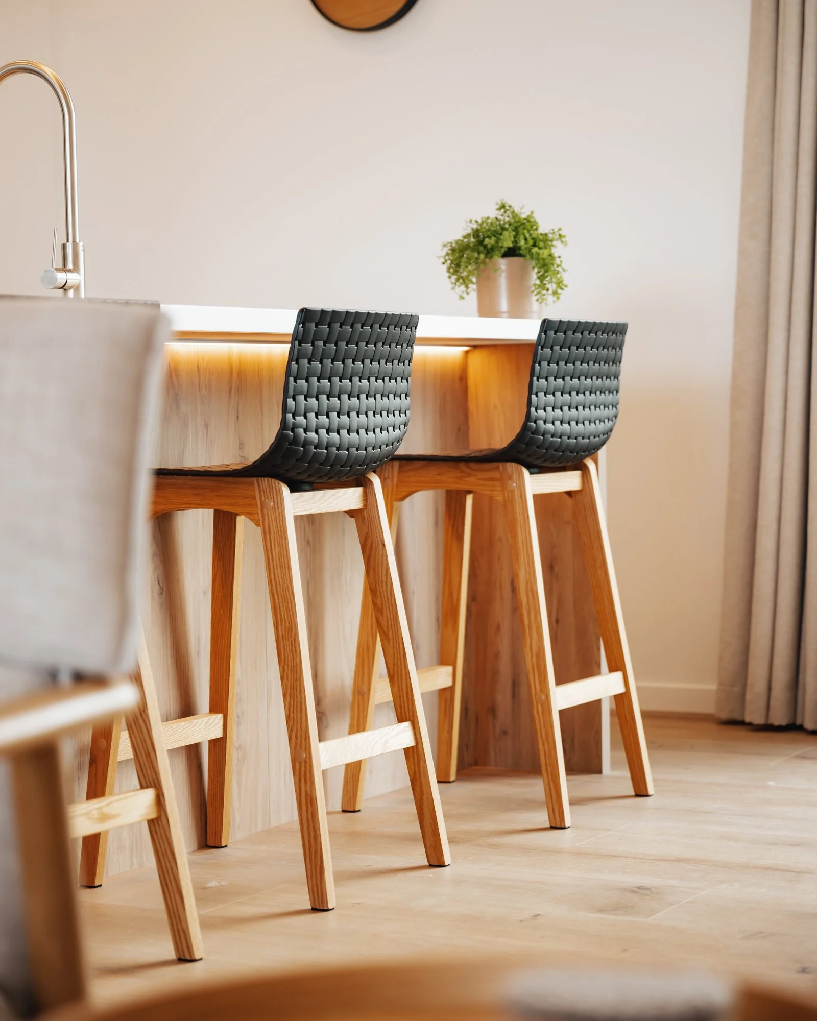 A kitchen island with two black woven chairs, a potted plant on the counter, and beige curtains on the window.
