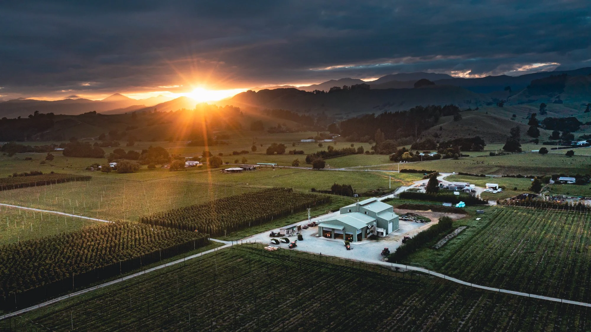 Aerial view of a rural landscape at sunset with fields, farm buildings, and distant mountains under a partly cloudy sky.