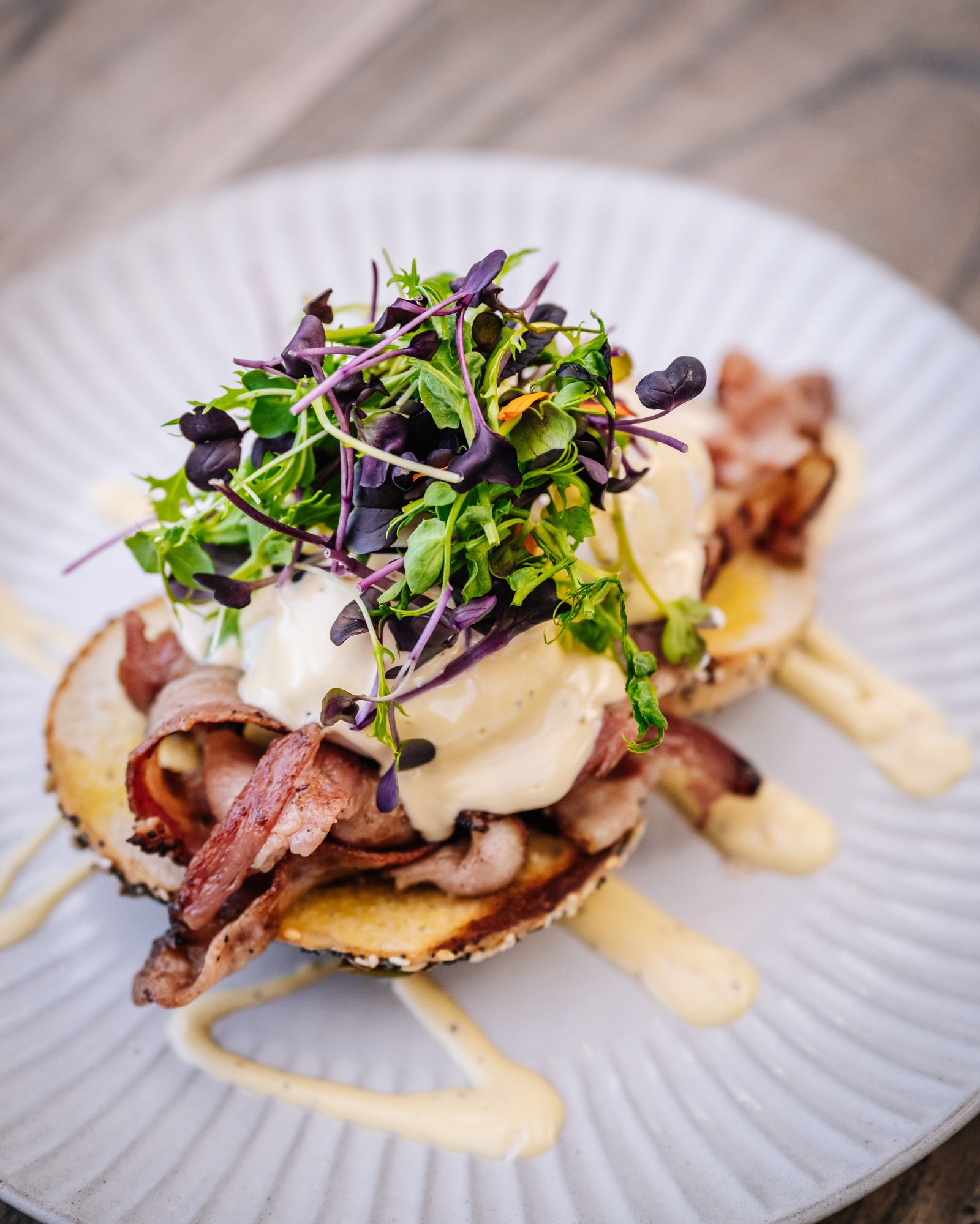 A gourmet dish featuring grilled bread topped with bacon, microgreens, a dollop of creamy sauce, and drizzled with a light sauce or aioli, served on a white textured plate.