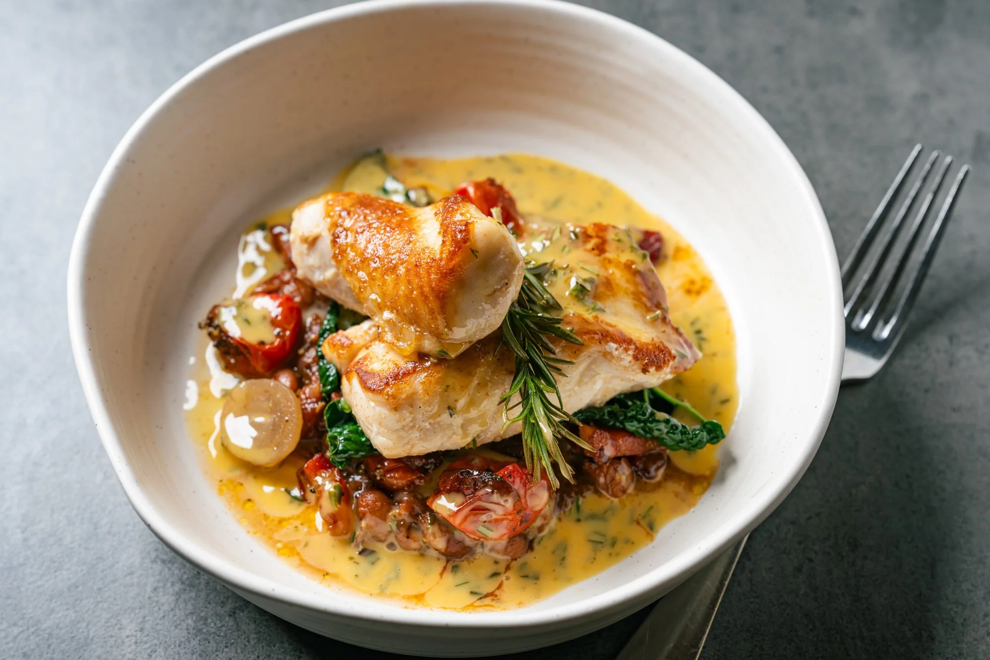 A white bowl containing cooked fish fillet topped with a sprig of rosemary, served with a yellow sauce, cherry tomatoes, capers, and greens.