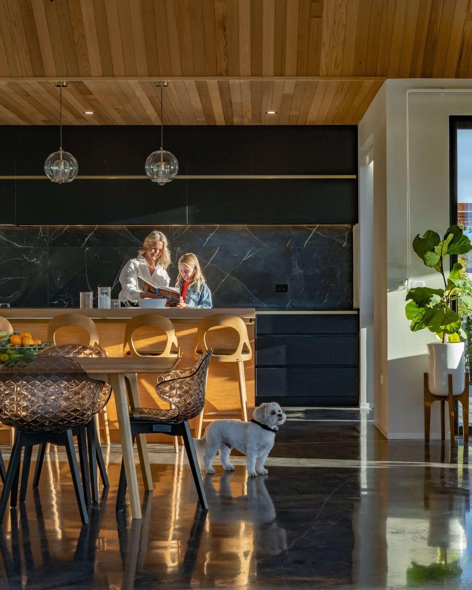 A modern kitchen with a wooden ceiling, black and marble backsplash, and a large window. A woman and a girl are at the kitchen counter looking at a book. A white dog with a black collar stands on the shiny marble floor near the table, which has chair