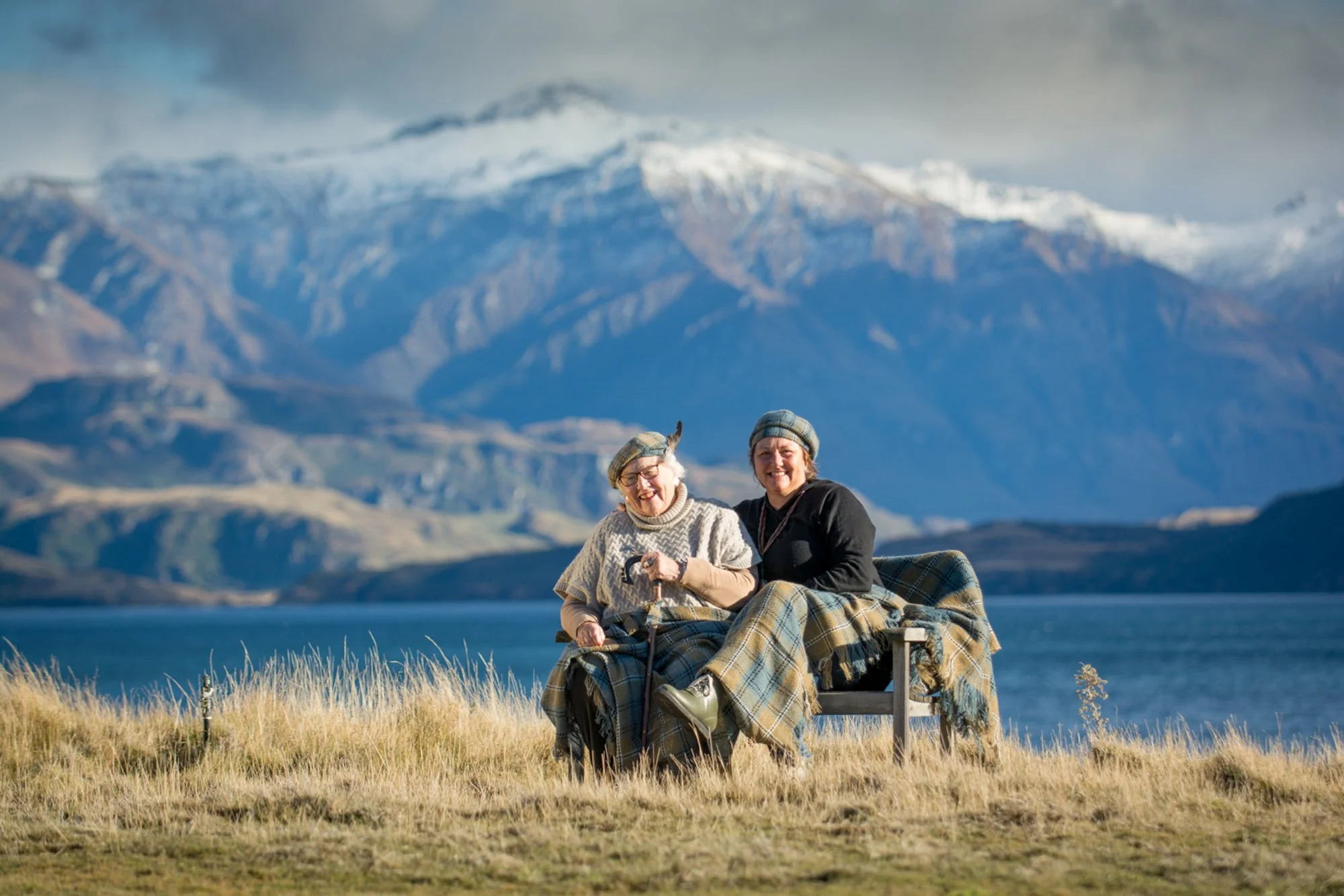 Two women sitting on a bench with a lake, mountains, and snow in the background, dressed warmly and smiling.