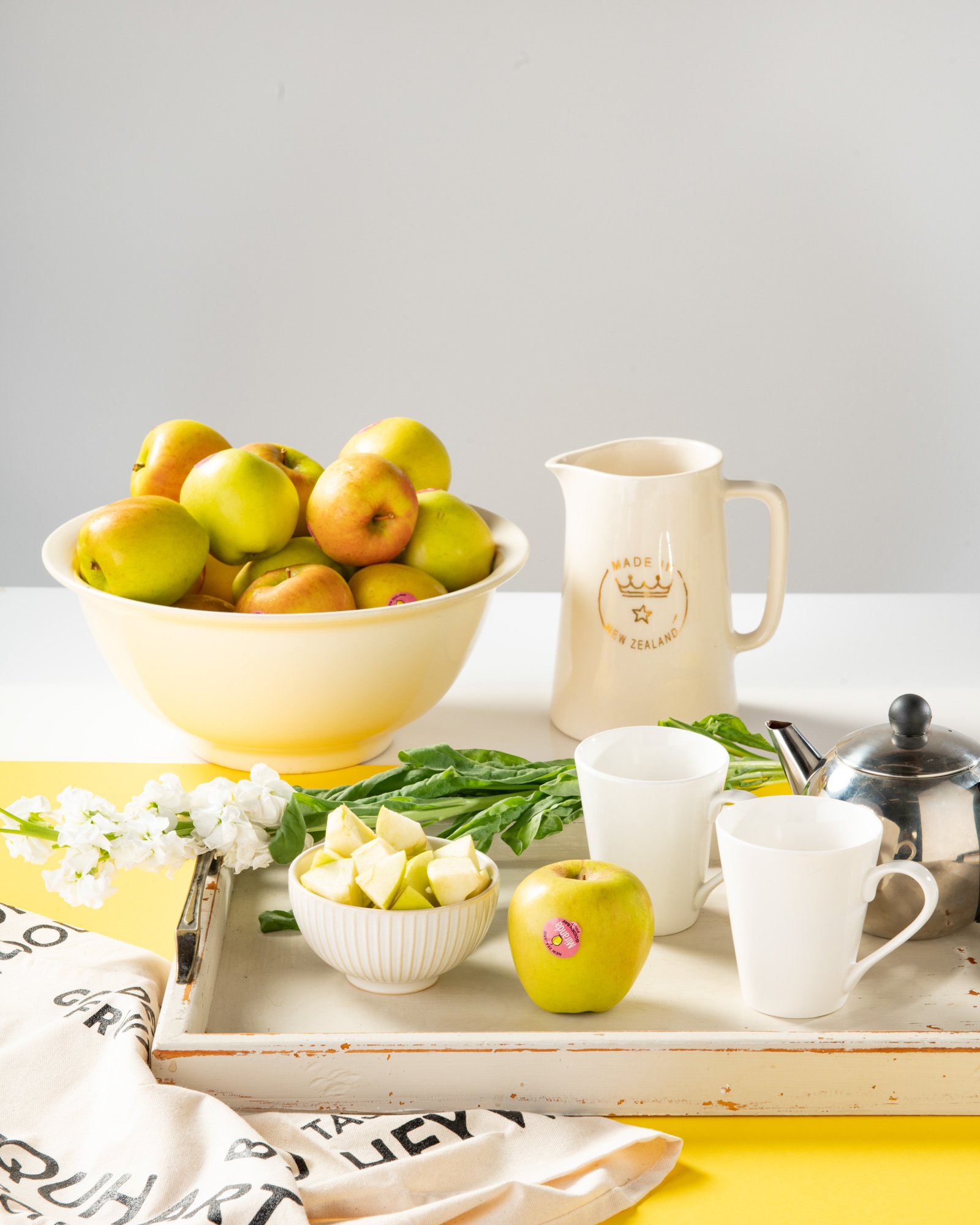 A still life of a bowl of apples, a white bowl of sliced apples, a pitcher with a 'Made in New Zealand' label, two white cups, a stainless steel teapot, fresh green leaves, and a white cloth with black writing, all arranged on a white and yellow surf