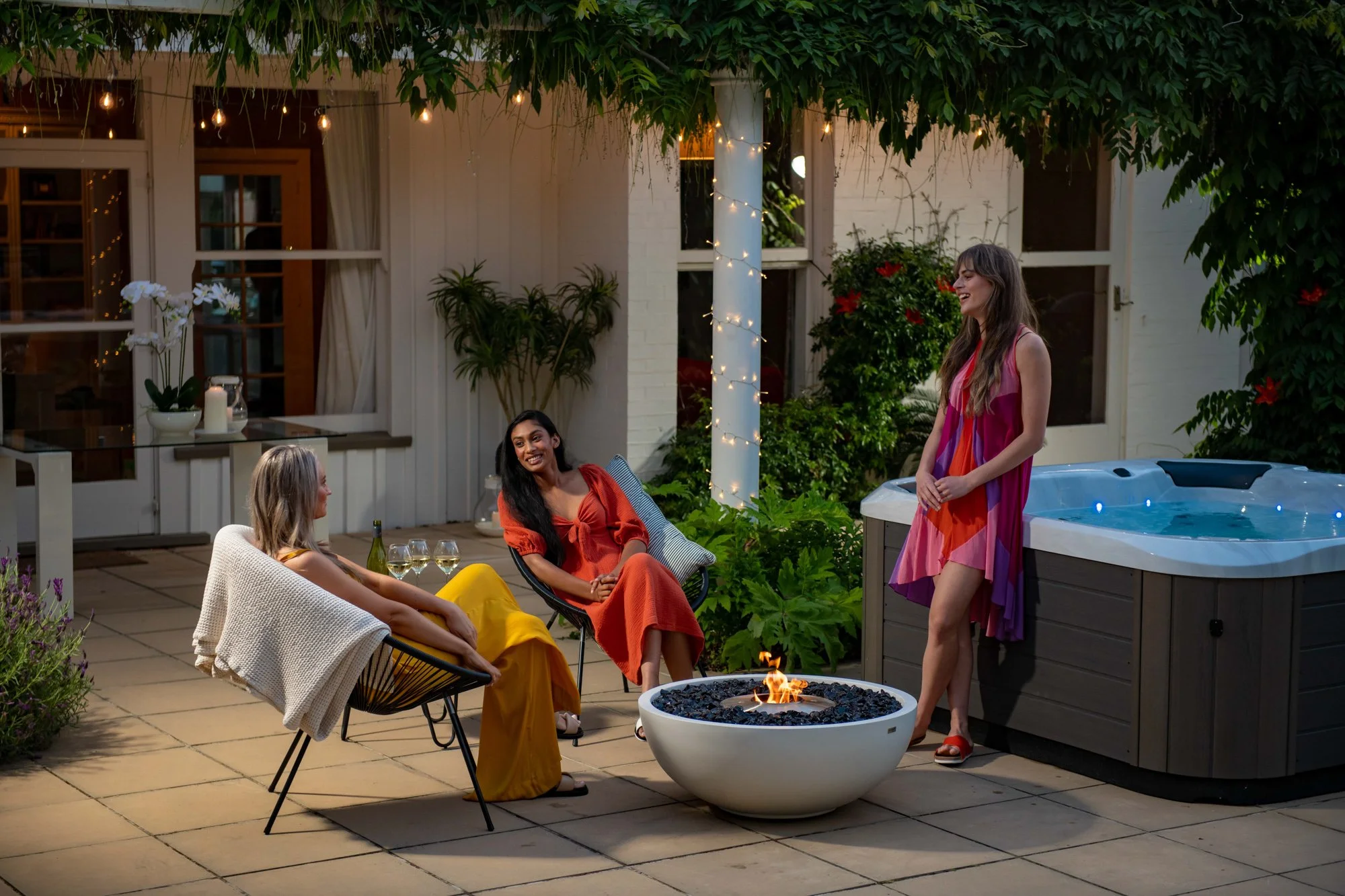 Four women enjoying an outdoor evening gathering on a patio, sitting by a fire pit, with one standing near a hot tub, in a setting decorated with string lights and lush greenery.