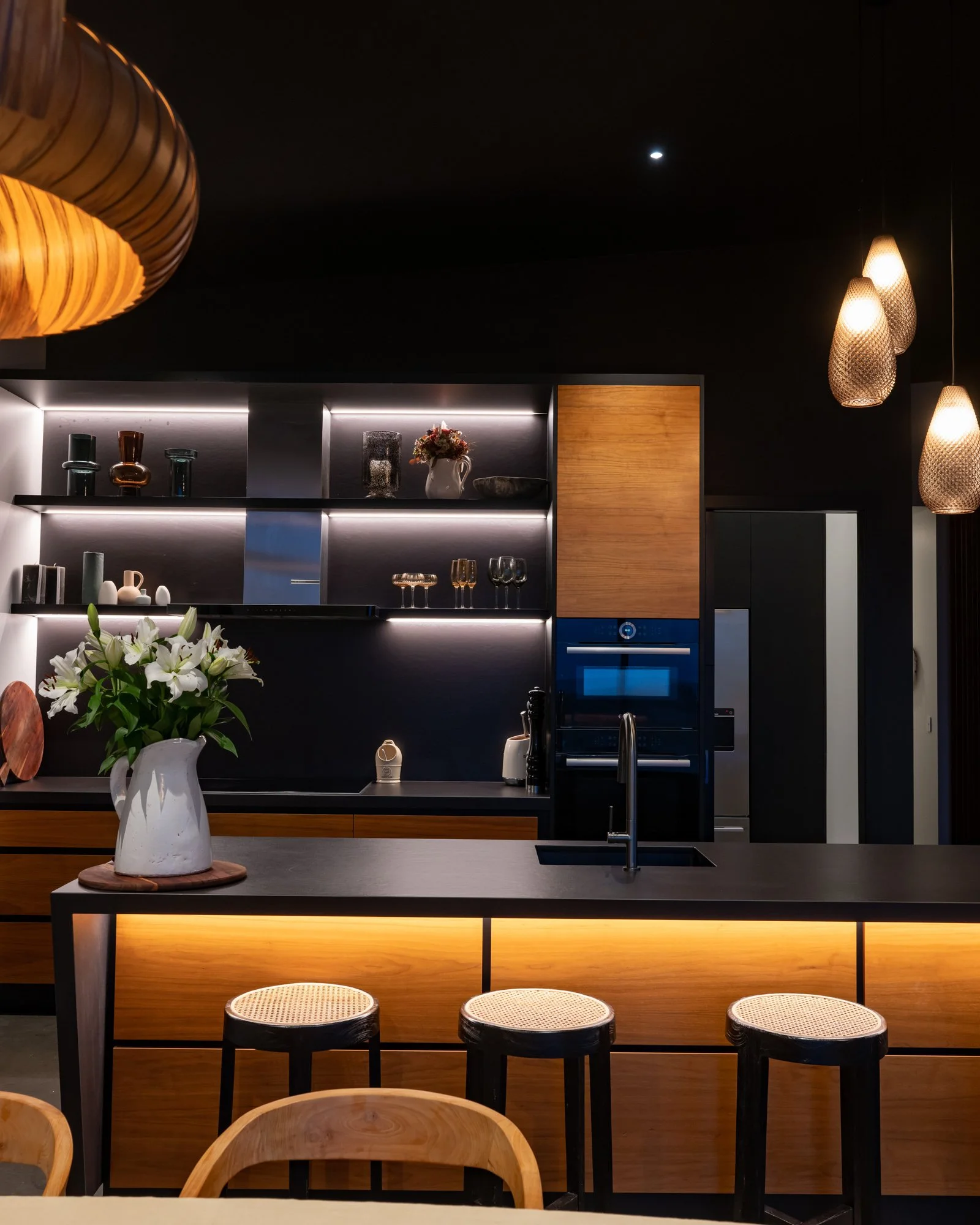 Modern kitchen with black and wooden cabinetry, illuminated open shelves with glassware and decorative items, a kitchen island with a vase of white lilies, and contemporary hanging pendant lights.