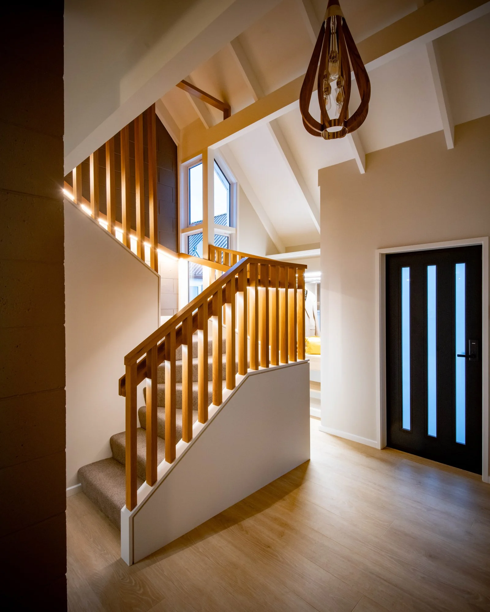 Modern entryway with a wooden staircase, large windows, and a dark front door with vertical glass panels, illuminated by natural and artificial light.