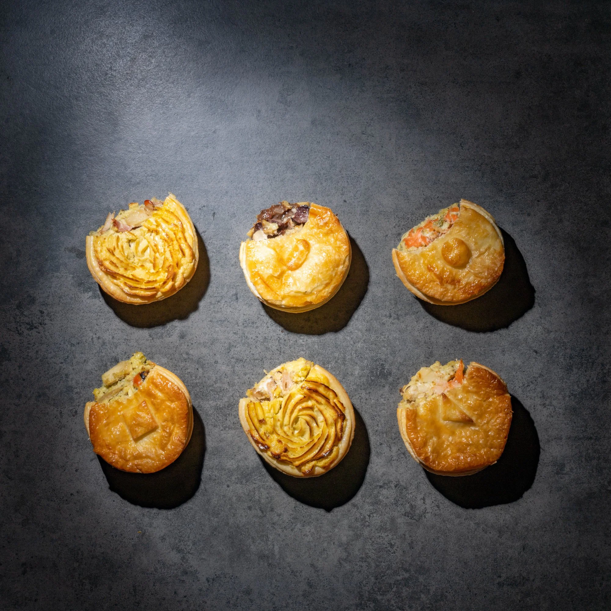 Six assorted mini pot pies on a dark surface, arranged in two rows of three. Each pie has different toppings and fillings visible at the edges.