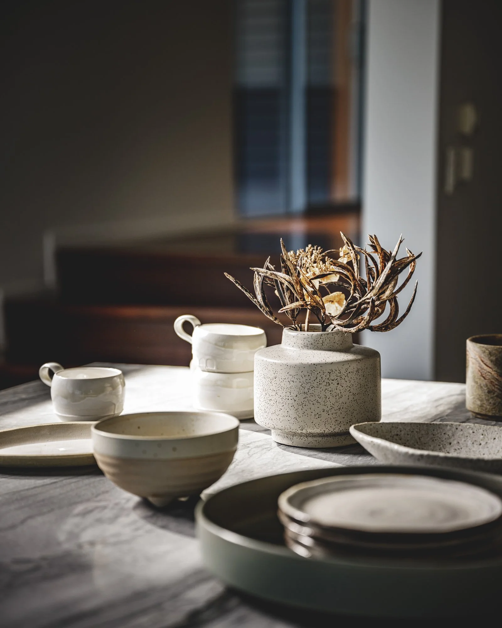 Ceramic bowls, plates, cups, and a vase with dried plant on a marble table, sunlight casting shadows.
