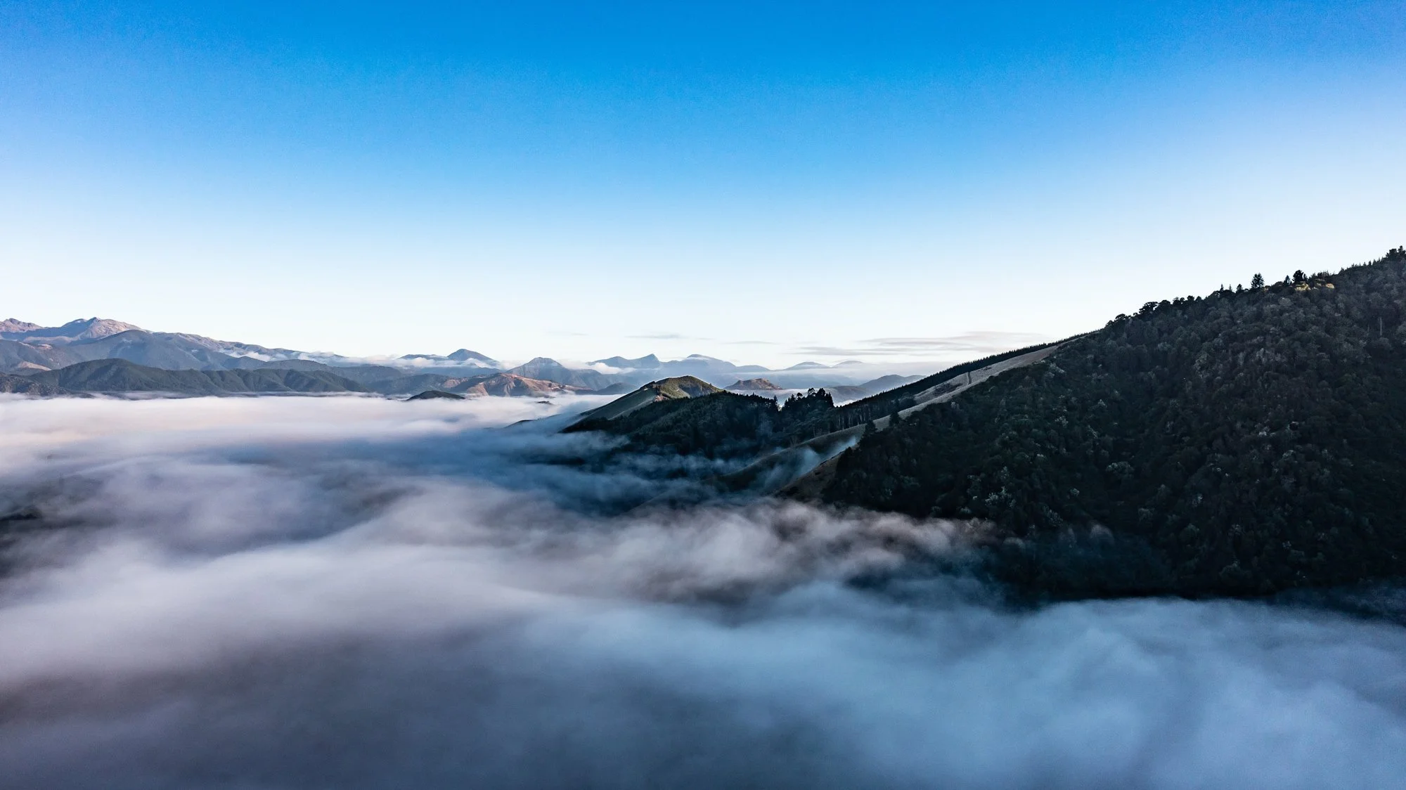 Mountain range with rolling peaks partially covered in clouds under a clear blue sky.