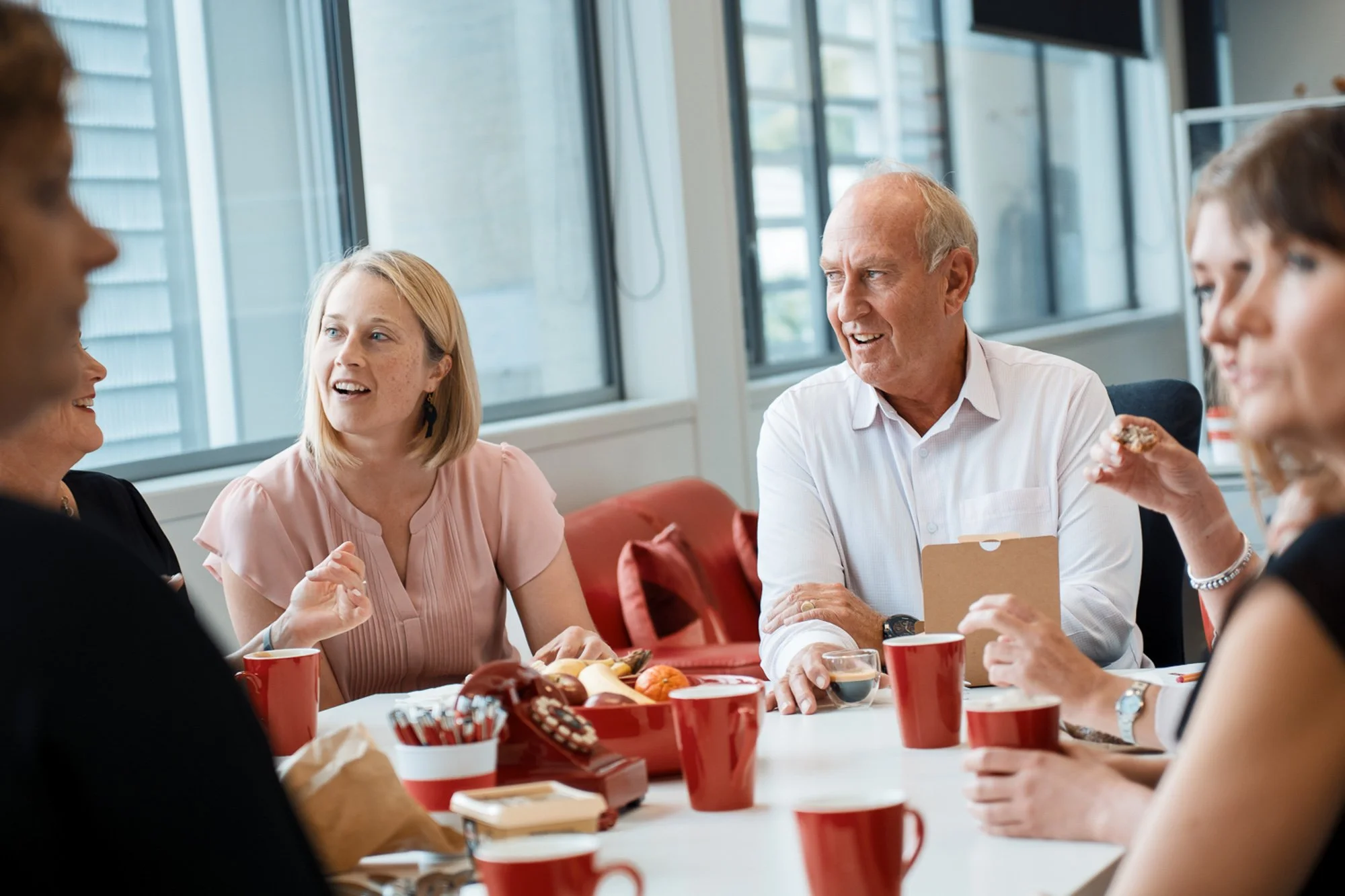 Group of six people having a discussion at a table with red cups and holiday decorations in a bright office space.