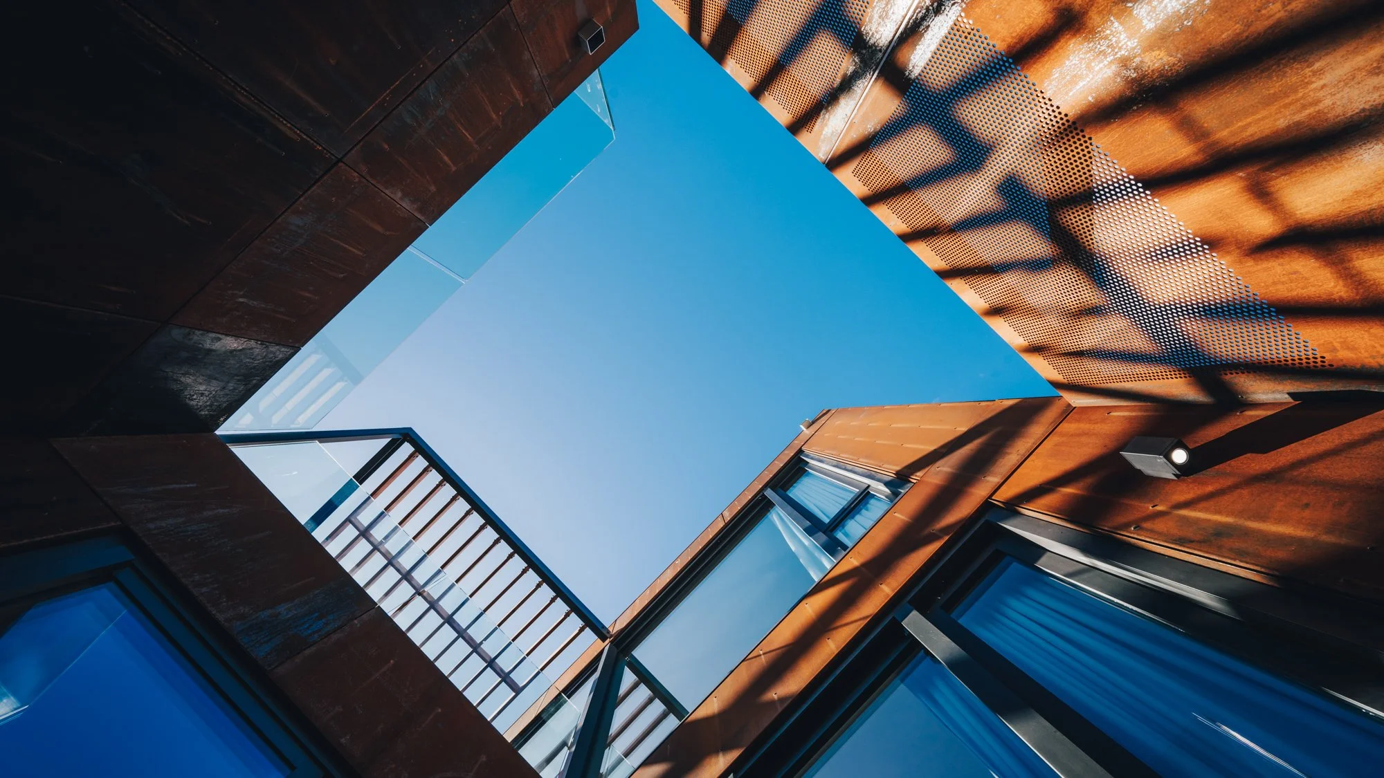 View looking up at a modern building courtyard with a clear blue sky above, showing metal and glass exterior walls with shadows cast by a staircase railing.