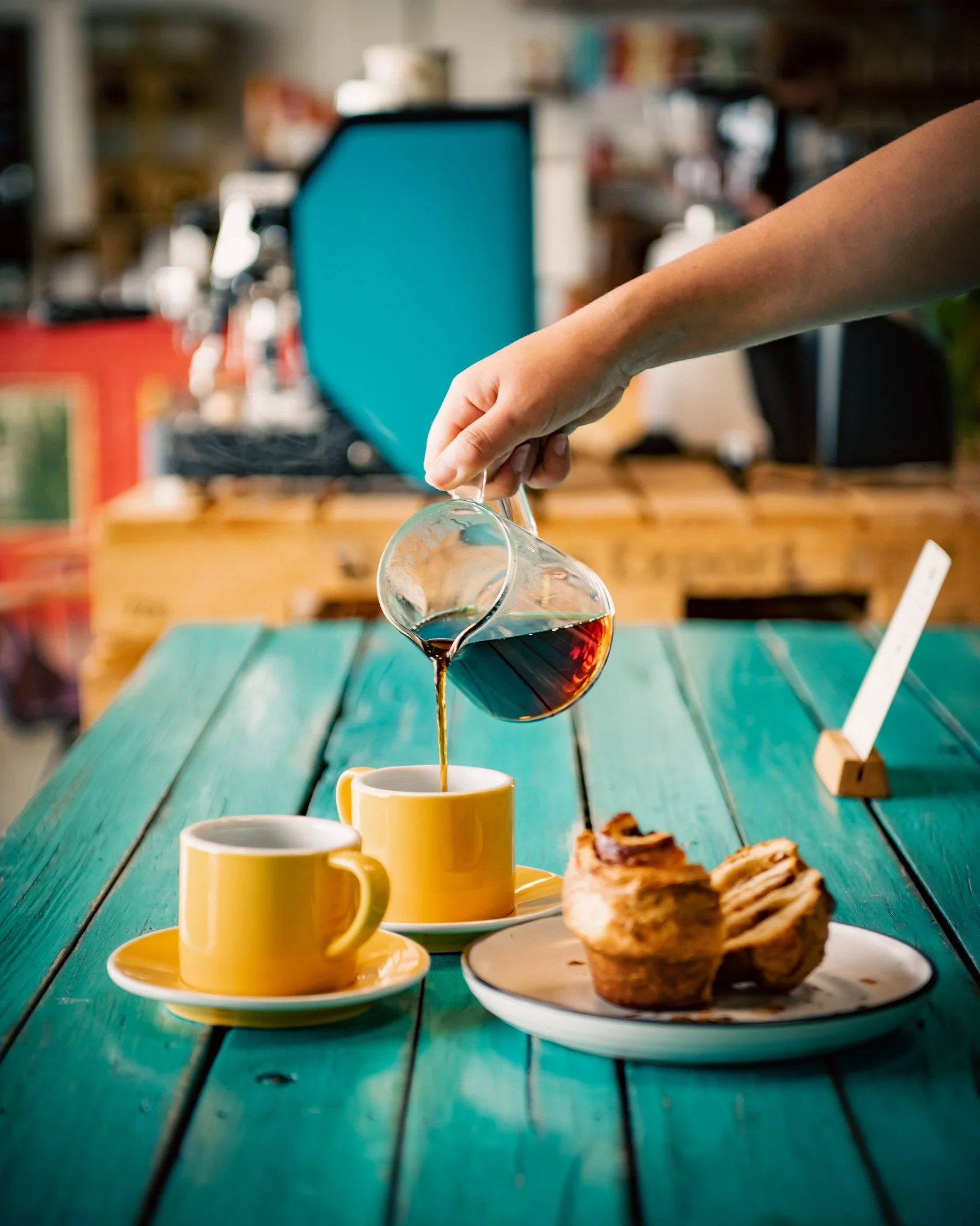 Person pouring coffee from a glass carafe into a yellow mug on a green wooden table next to pastries and another yellow mug, in a cozy cafe setting.