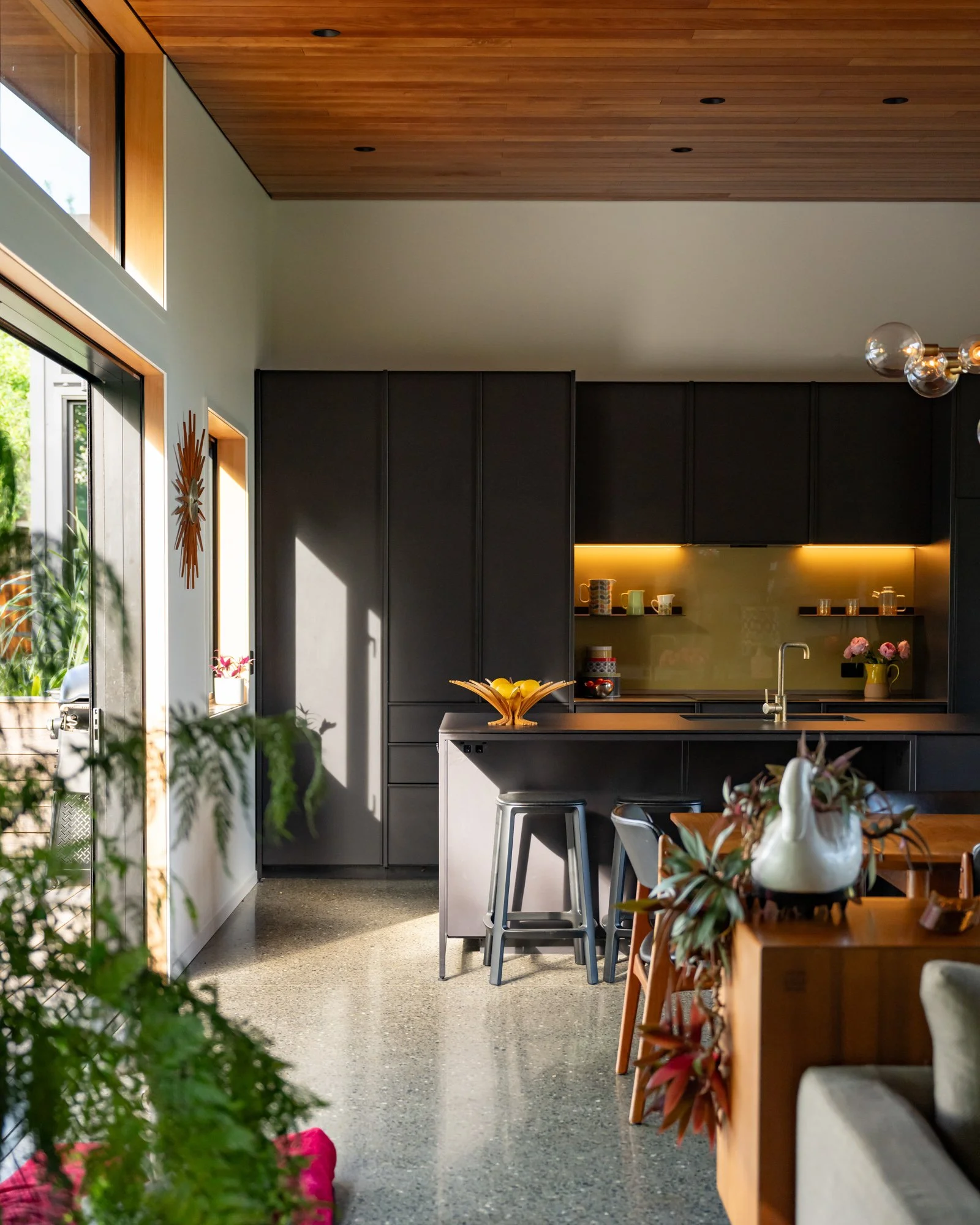 Modern kitchen with black cabinets, wooden ceiling, and large windows letting in natural light. Decor includes plants, a bowl of lemons, and flowers on the table.