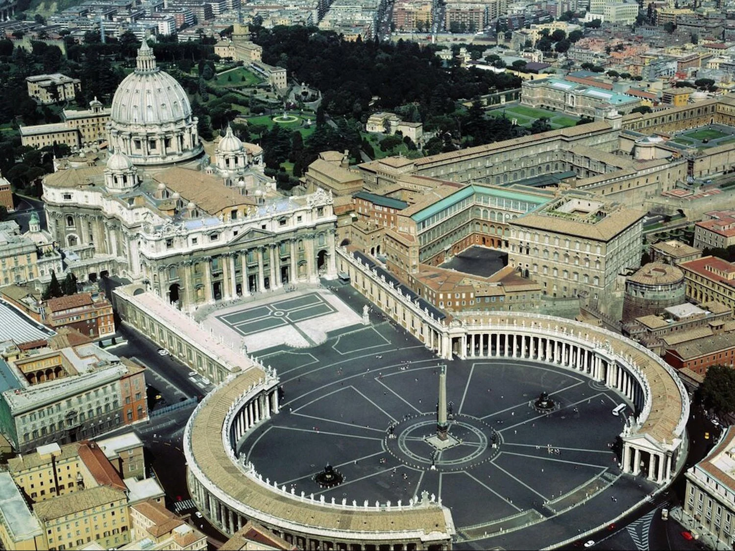 An aerial view of Saint Peter's Square that includes Saint Peter's Basilica, an Egyptian obelisk, the colonnade the embraces the obelisk, and some of the surrounding area.