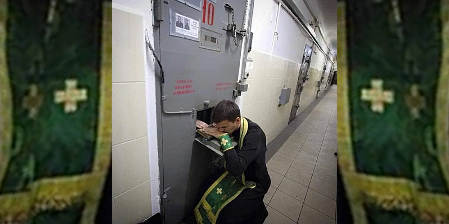 A priest is hearing confession from a prisoner in solitary confinement. The prisoner is behind a gray metal door with a small door some three feet above the floor. Only his right hand shows as the priest is squatting to listen through that opening.