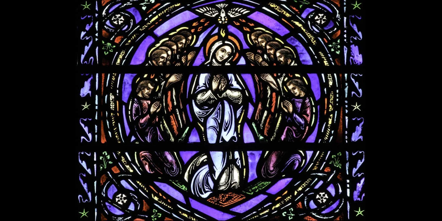 Stained glass window depicting Pentecost.  In the center is the Blessed Mother in white.  Above her is the Holy Spirit depicted as a dove from whom come tongues of fire.  The Eleven Apostles form a circle around the Blessed Mother.