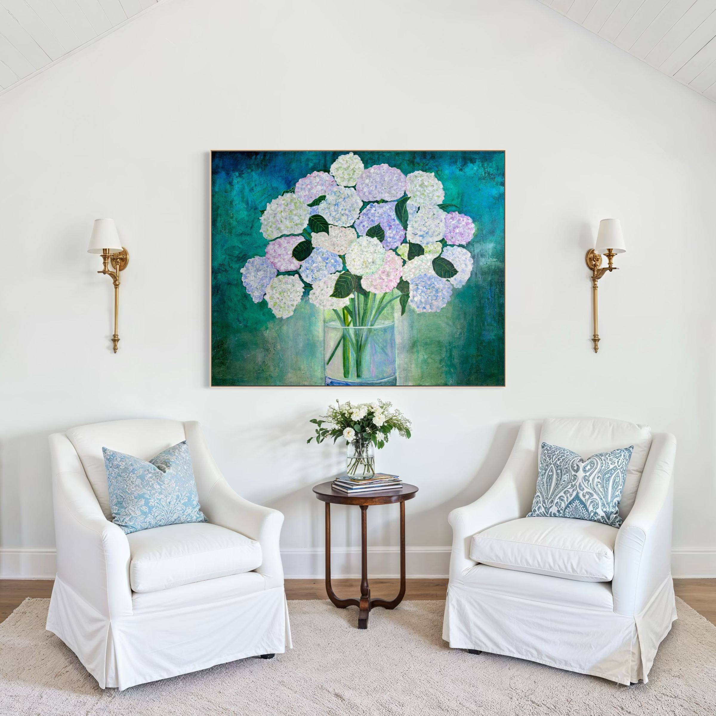 Living room with two white armchairs, floral pillows, a small wooden table with a vase of white flowers and stacks of magazines, large floral painting, and wall lamps.