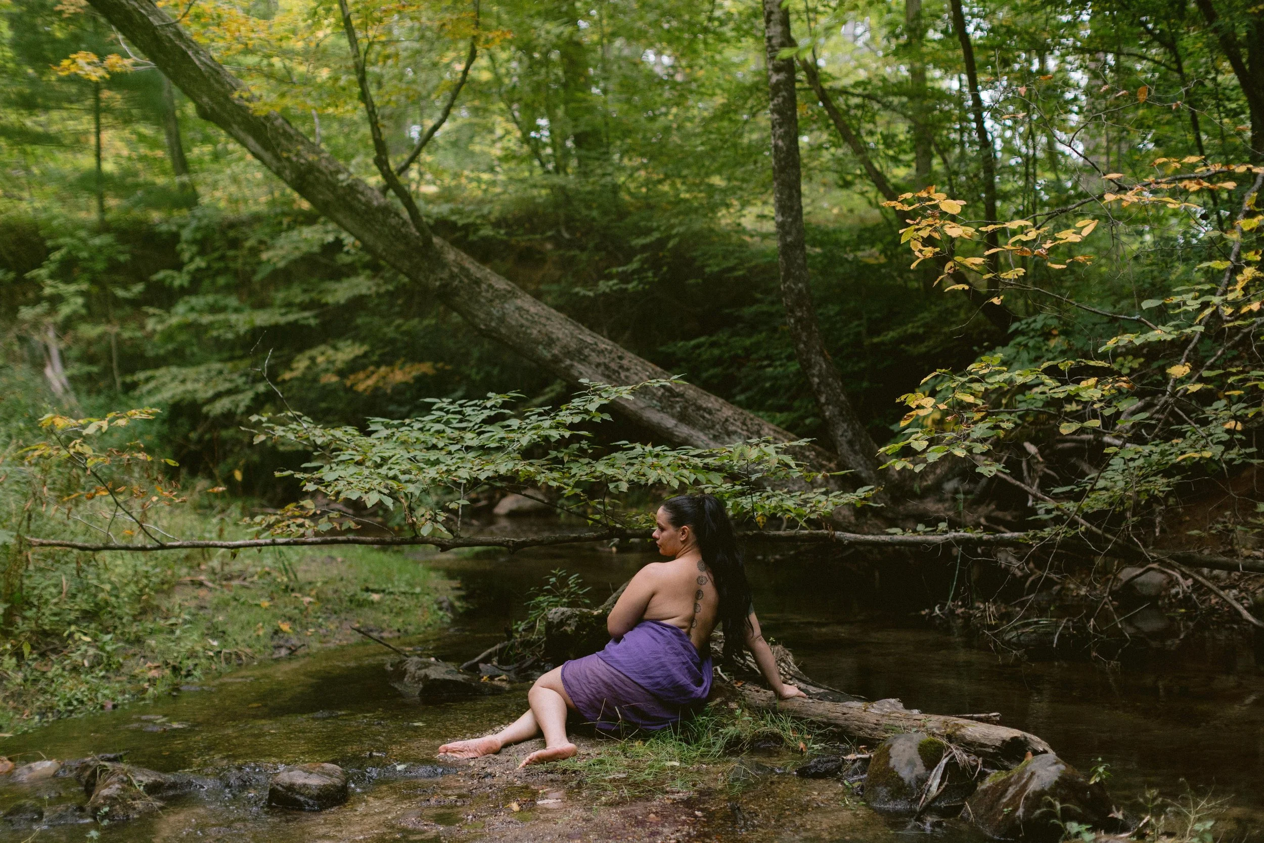 Woman with long dark hair and tattoos, wrapped in a purple towel, sitting by a creek in a lush green forest with trees and fallen branches.