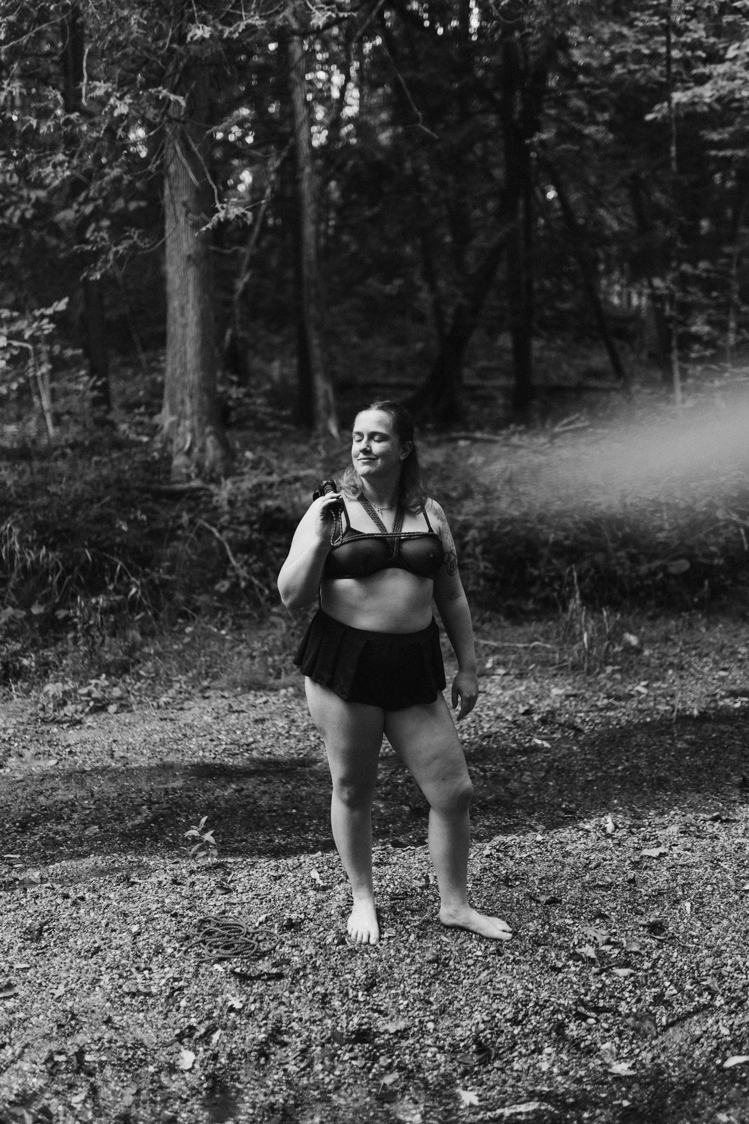 A woman with long hair, wearing a black sports bra and a short skirt, stands barefoot on a dirt path in a forest, holding a camera and smiling with her eyes closed.