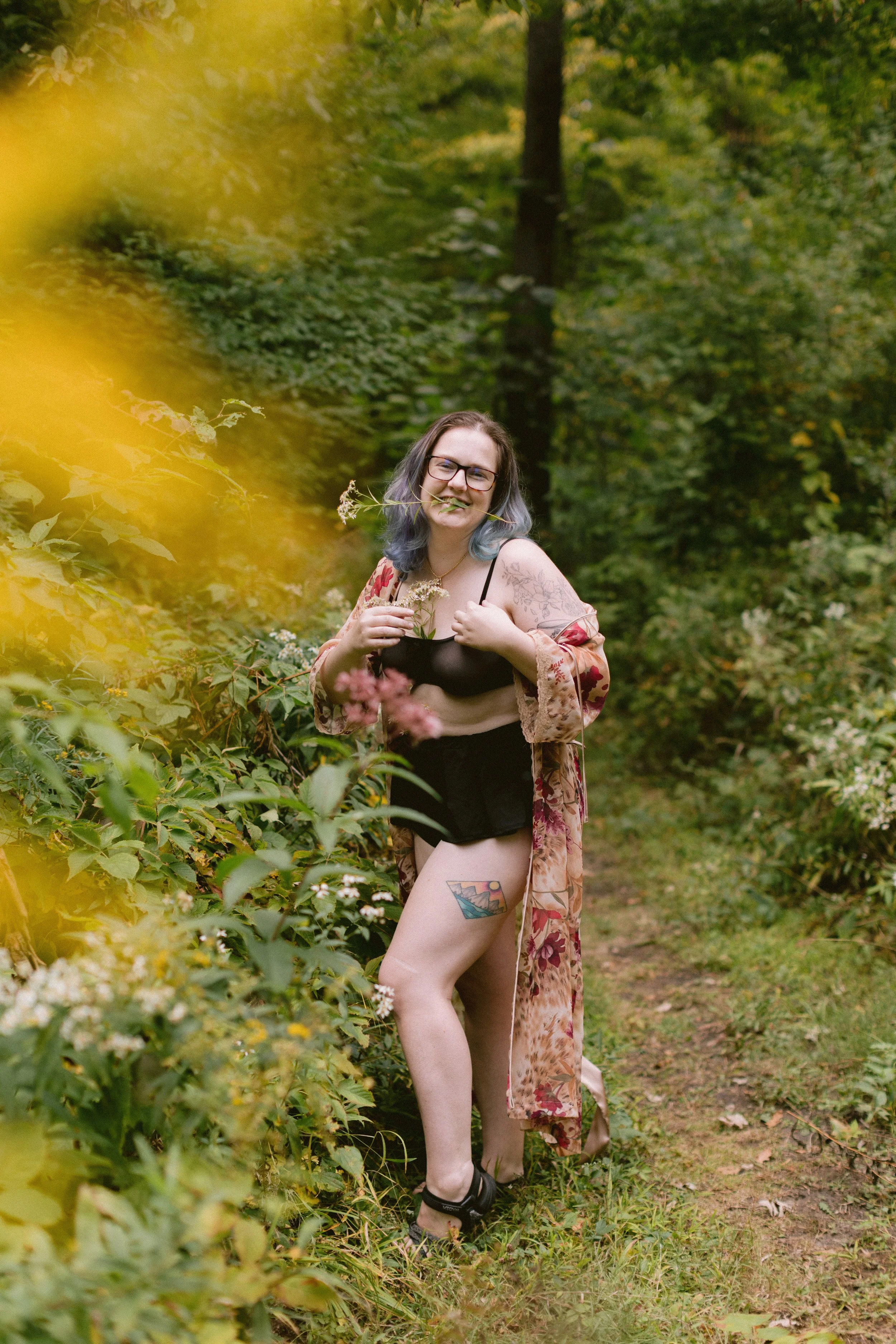 A woman with glasses and dyed hair smiling in a forest, wearing a black top, patterned kimono, and shorts, holding a plant stem in her mouth, with a colorful tattoo on her thigh.
