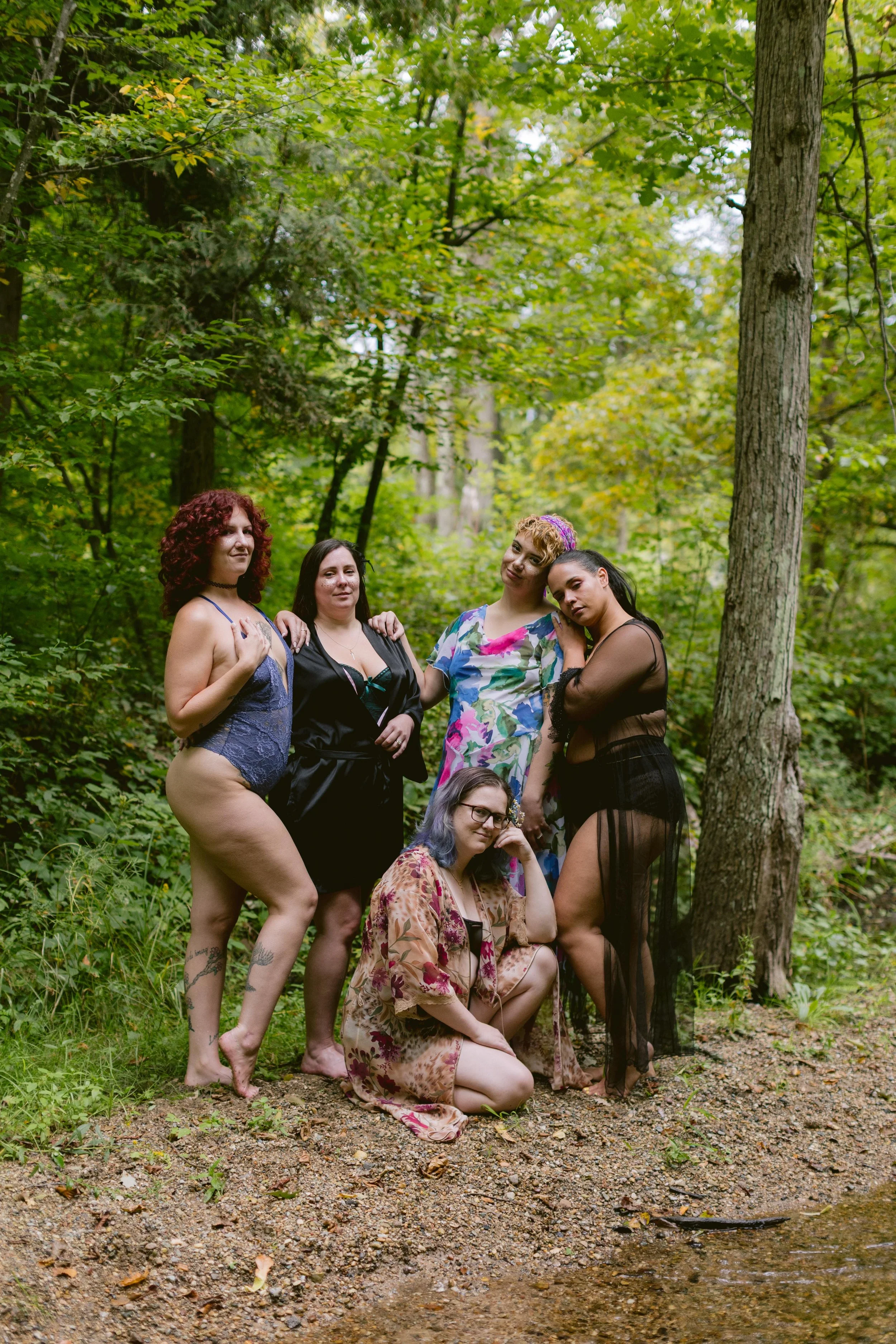 Group of six women standing and sitting in a wooded area with green trees and a small creek.