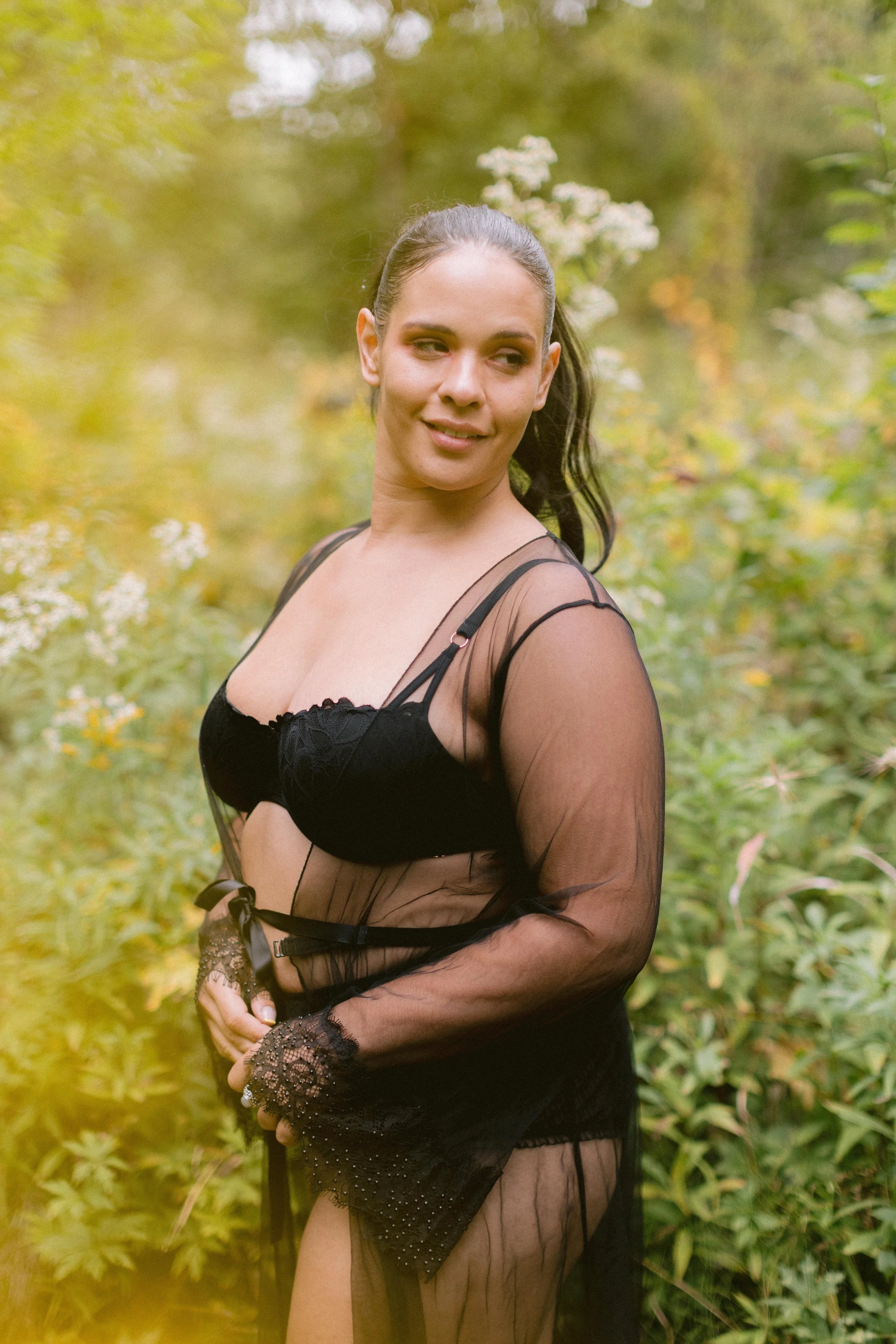 A woman in black lace lingerie and a sheer black robe stands outdoors among green foliage, with a slight smile and her hair in a ponytail.
