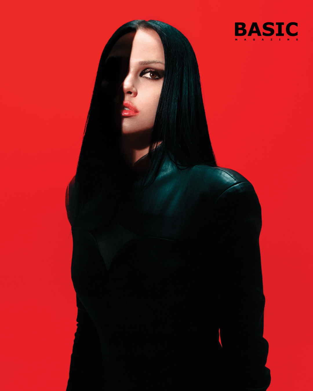 A woman with long black hair and fair skin, wearing a black leather jacket, looking at the camera with red lipstick on, against a solid red background. The text 'BASIC Magazine' is in the top right corner.
