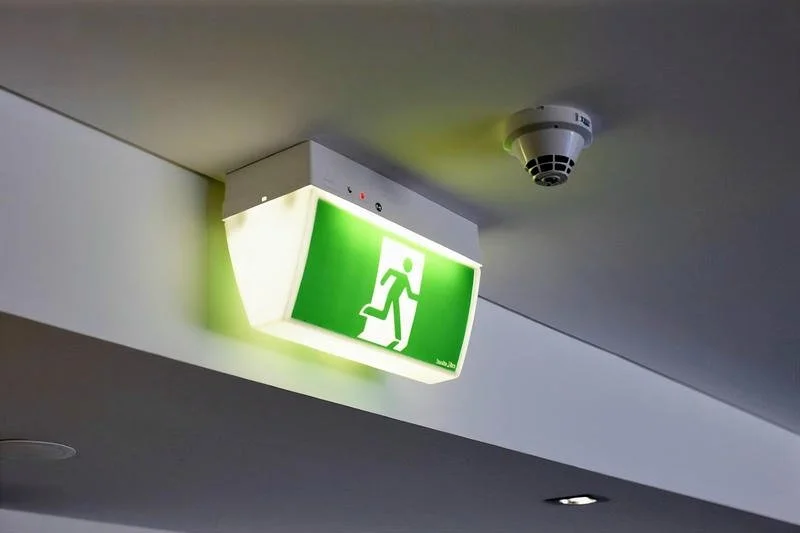 An illuminated green emergency exit sign with a white running person and an arrow pointing left, attached to a ceiling in a building.