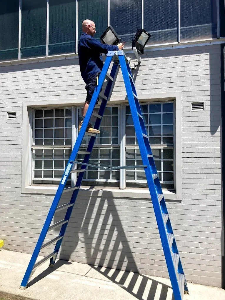 A man standing on a blue ladder working on two external flood lights attached to a building's wall during daytime.