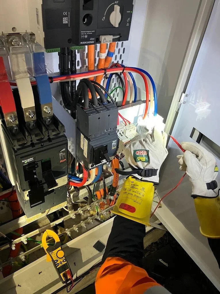 An electrician working on an electrical panel, handling wires with a yellow safety glove on the right hand. The panel contains circuit breakers, wires, and electrical components.
