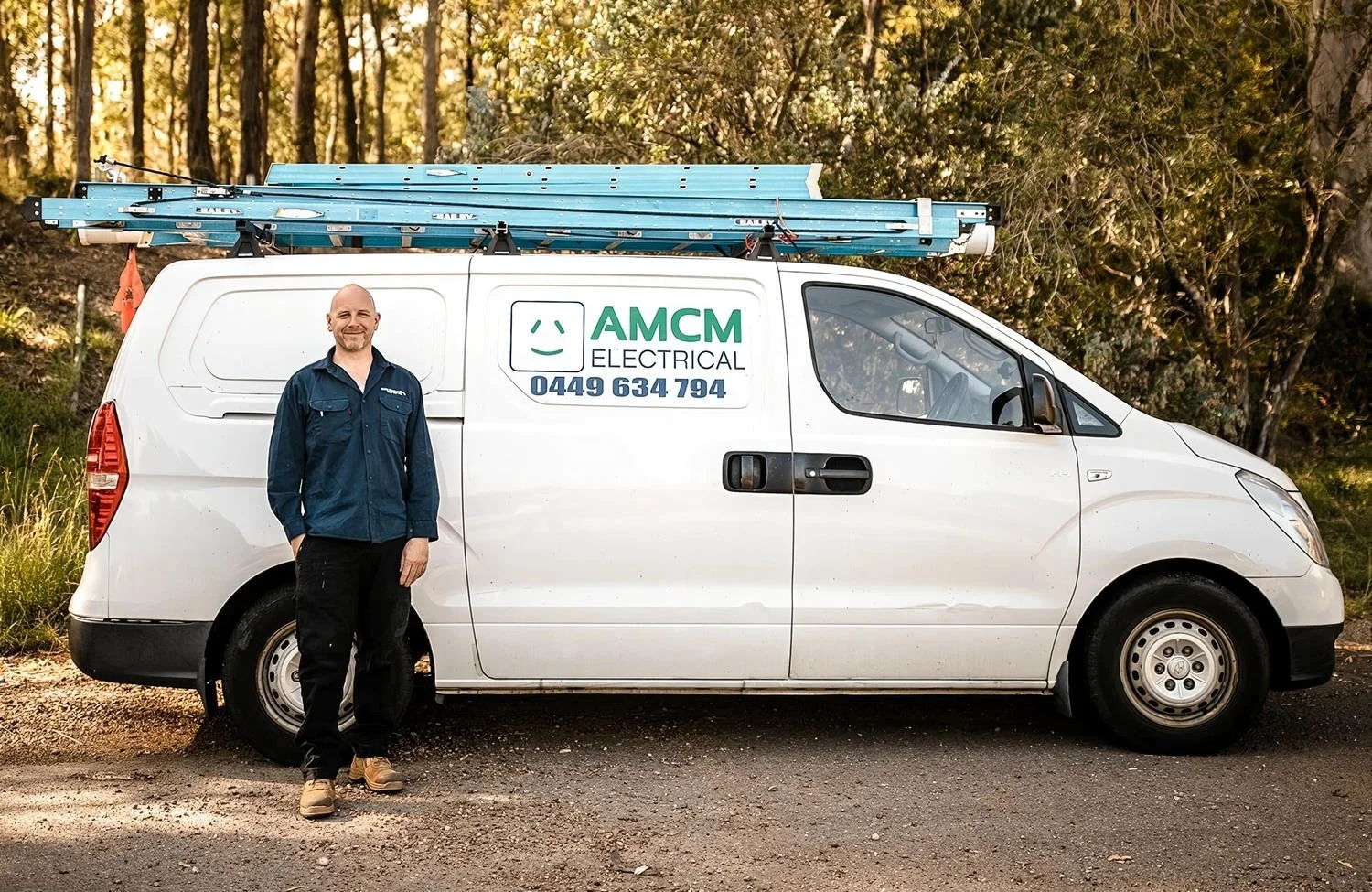 A smiling electrician in a navy blue shirt and black pants standing next to a white service van with a sign that reads 'AMCM Electrical' and a phone number, with a blue ladder on the roof, parked on a dirt road with trees in the background.