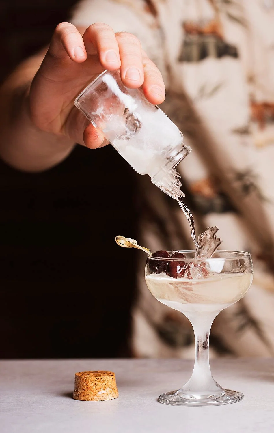 A bartender pouring a clear liquid into a cocktail glass with cherries and ice, with a cork on the table in front for a food and beverage photoshoot in Tulum . 