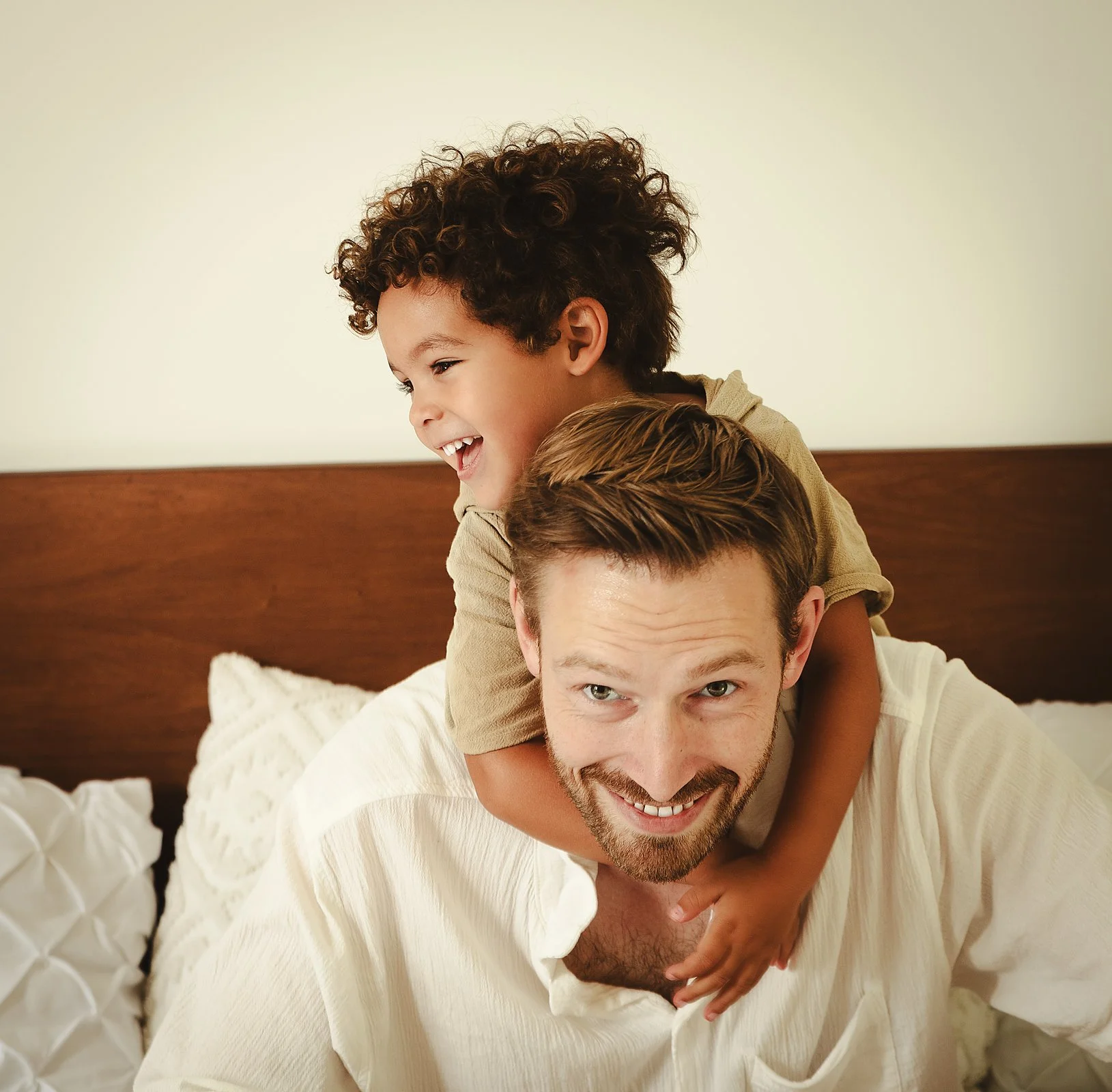 A father giving his young son a piggyback ride on a bed, both smiling and laughing in a family portrait in Tulum, Mexico.