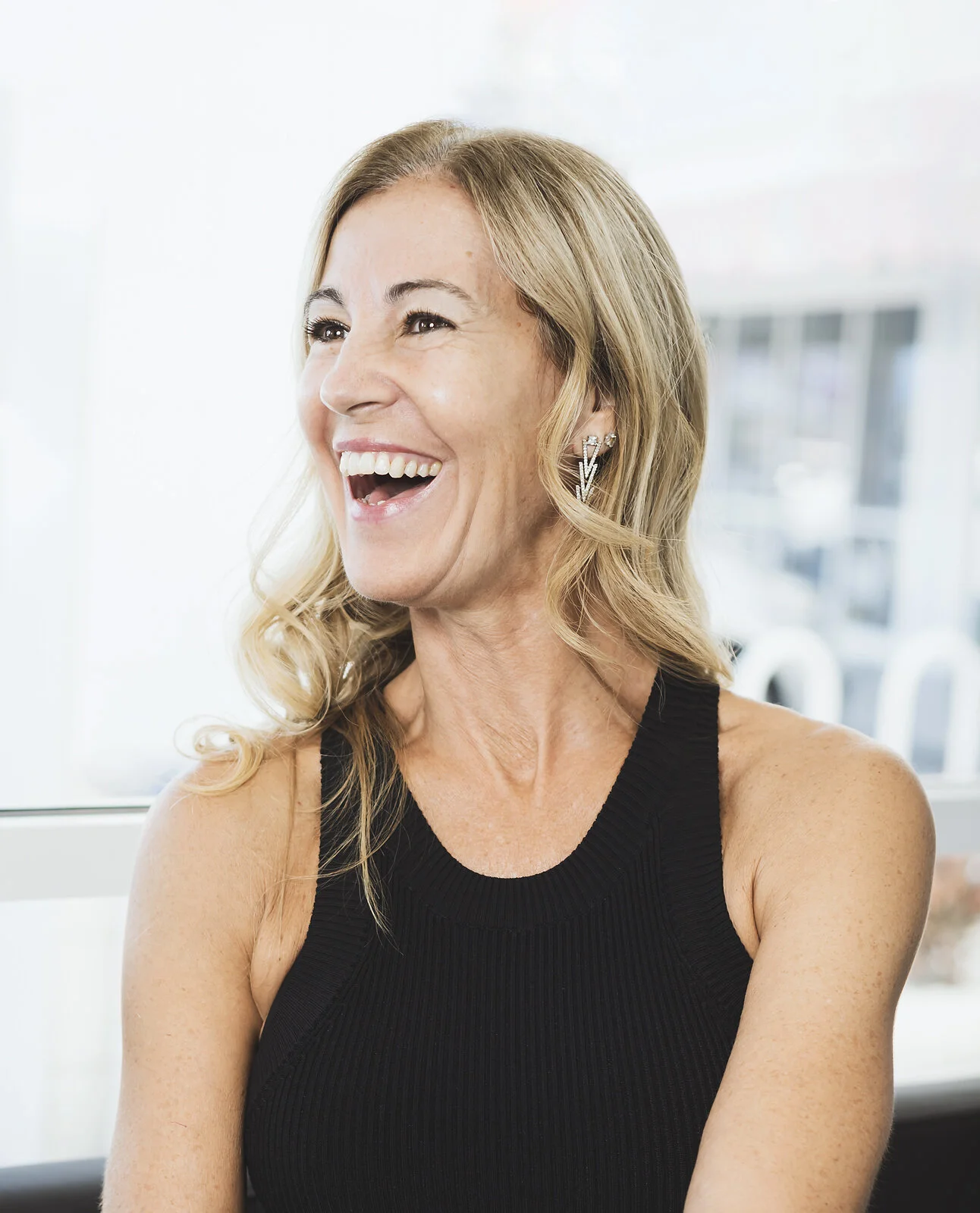 A headshot of a middle-aged woman with blonde wavy hair, smiling and wearing a black sleeveless top and butterfly-shaped earrings, indoor setting with bright natural light. Branding headshot. 
