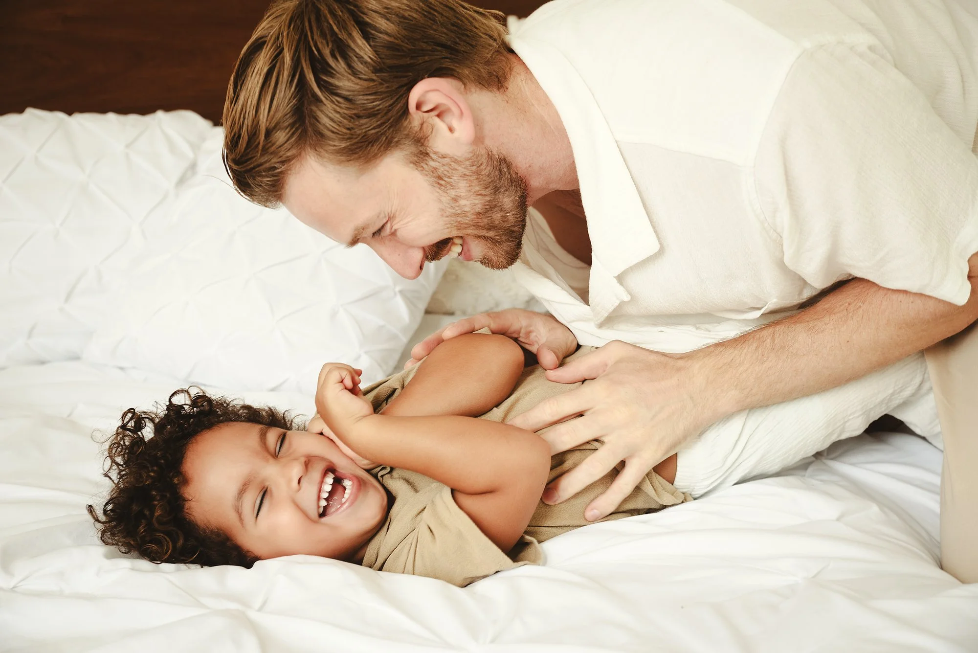 A man and son playing on a bed, smiling and playing together, with the man leaning over his son in a family portrait in Tulum, Mexico.