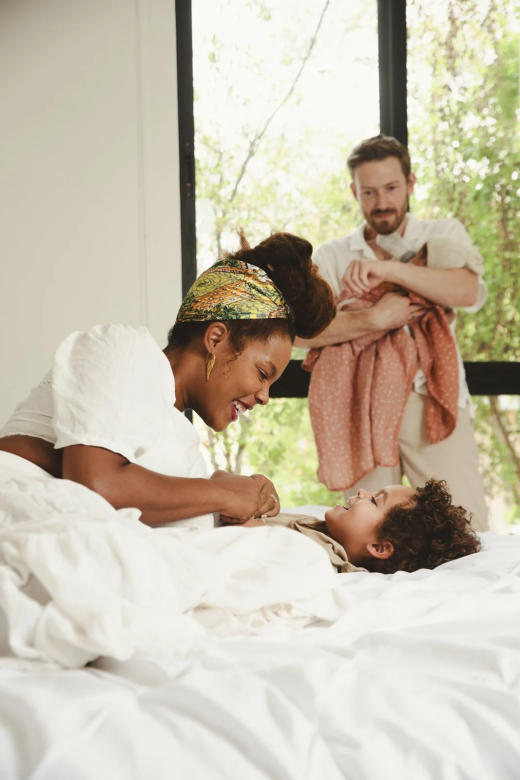A woman and a young boy lying on a bed, smiling at each other while the woman plays with the boy's hands. In the background, a man is holding a small child, standing near a window with green trees outside in this family portrait in Tulum, Mexico.