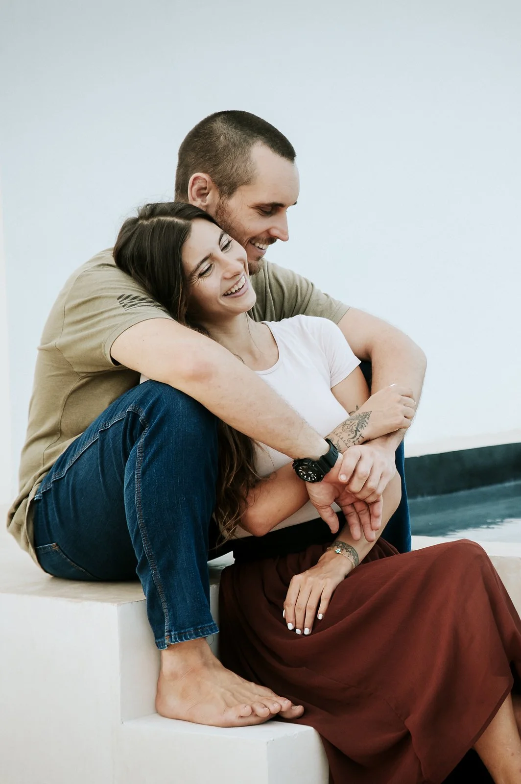 A happy couple sitting on a ledge, embracing and smiling, with a plain wall in the background in Tulum, Mexico.