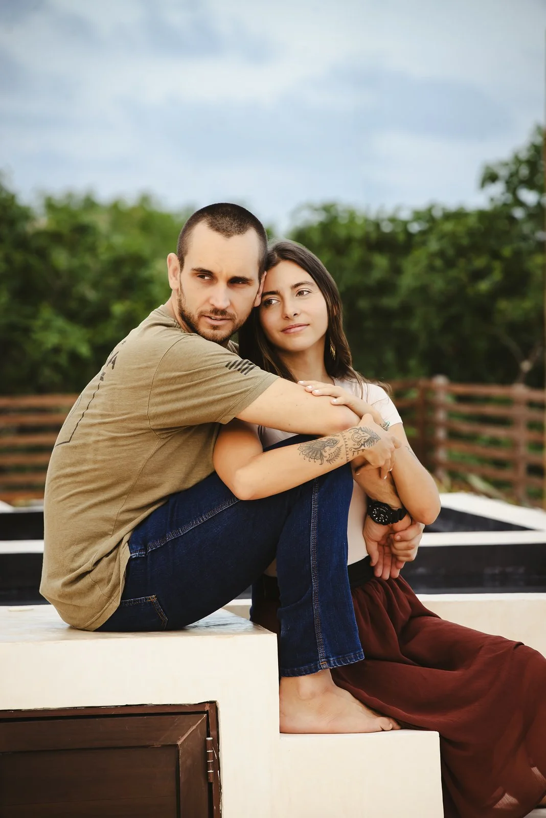 A man and woman sitting outdoors on a ledge, with the man hugging the woman and both looking contemplative. The background is filled with greenery and an overcast sky in a couples photoshoot in Tulum, Mexico.