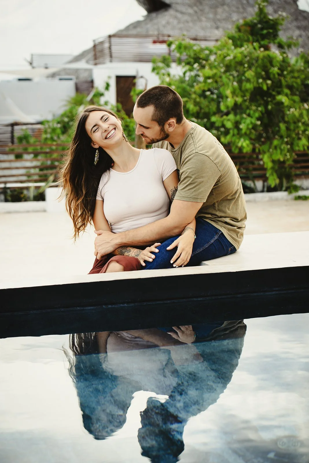 A couple sitting at the edge of a pool, smiling and embracing each other, with a rocky hill and greenery in the background, and their reflection visible in the water in Tulum, Mexico.