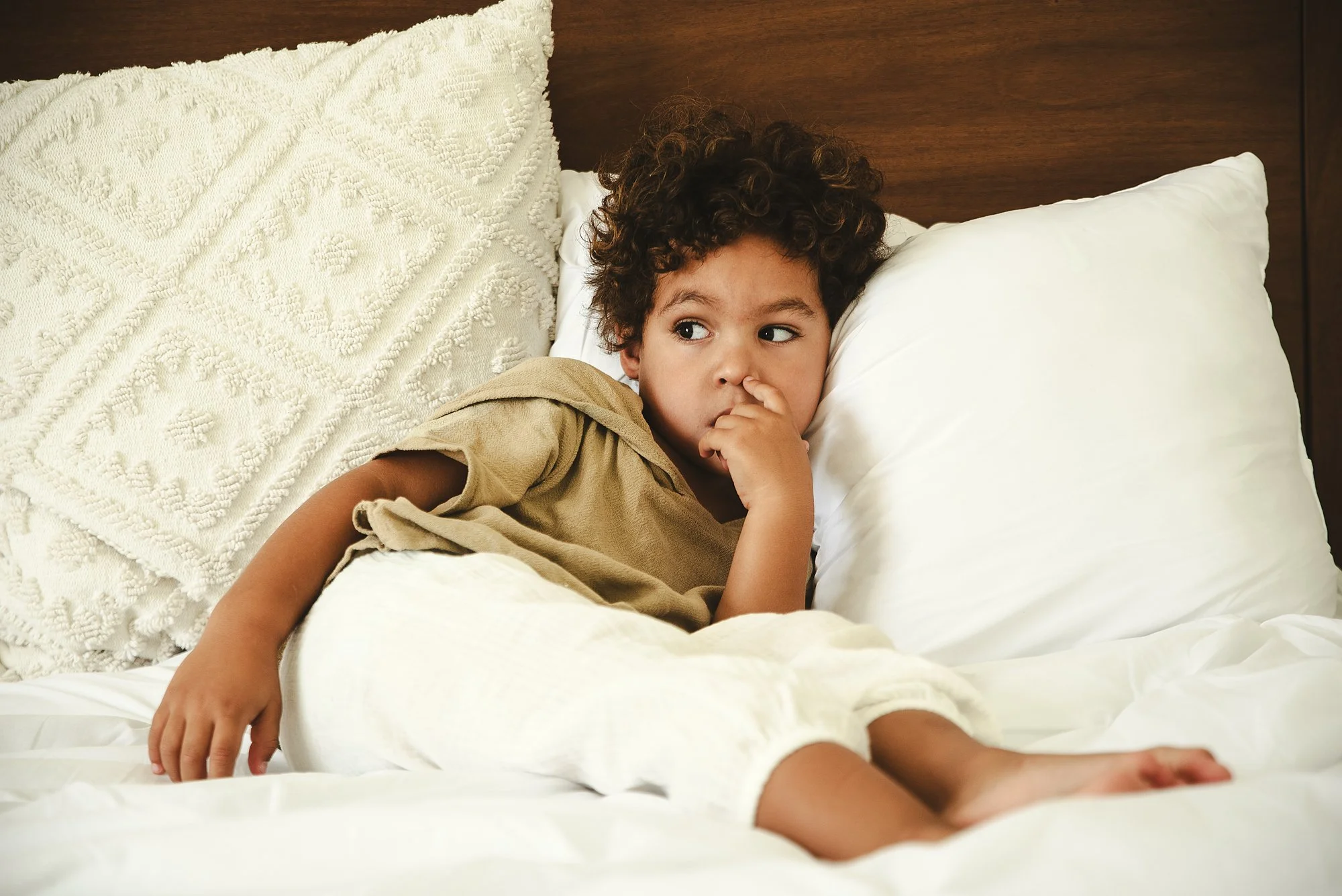 A young boy with curly hair lying on a bed, wearing a tan shirt, with one hand near his face and the other resting on the bed, looking off to the side in a family photoshoot in Tulum, Mexico.
