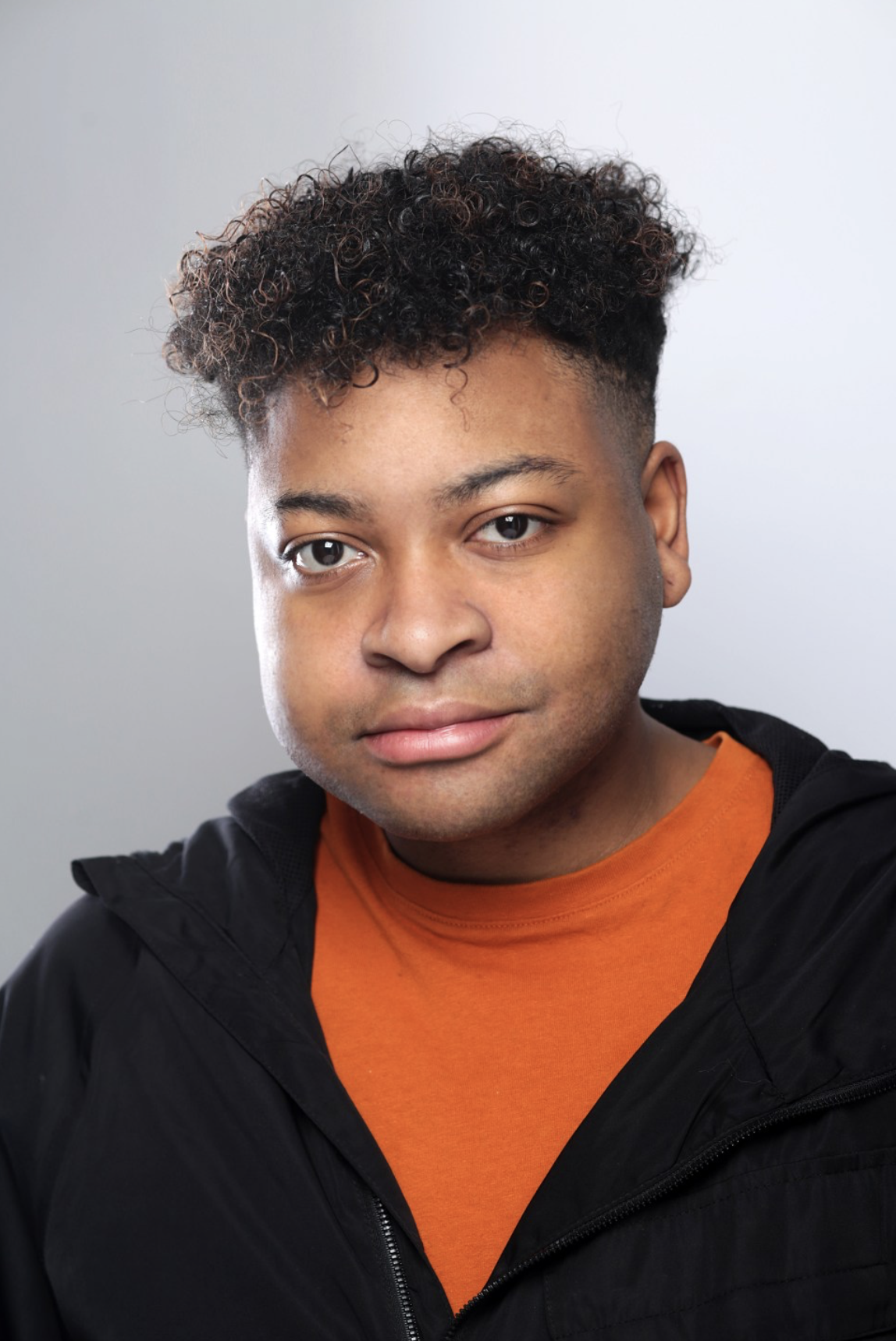 Headshot of actor Justin Brady. Justin is a black male between the ages of 17-27. He has black hair and is wearing an orange t-shirt with a black hoodie over