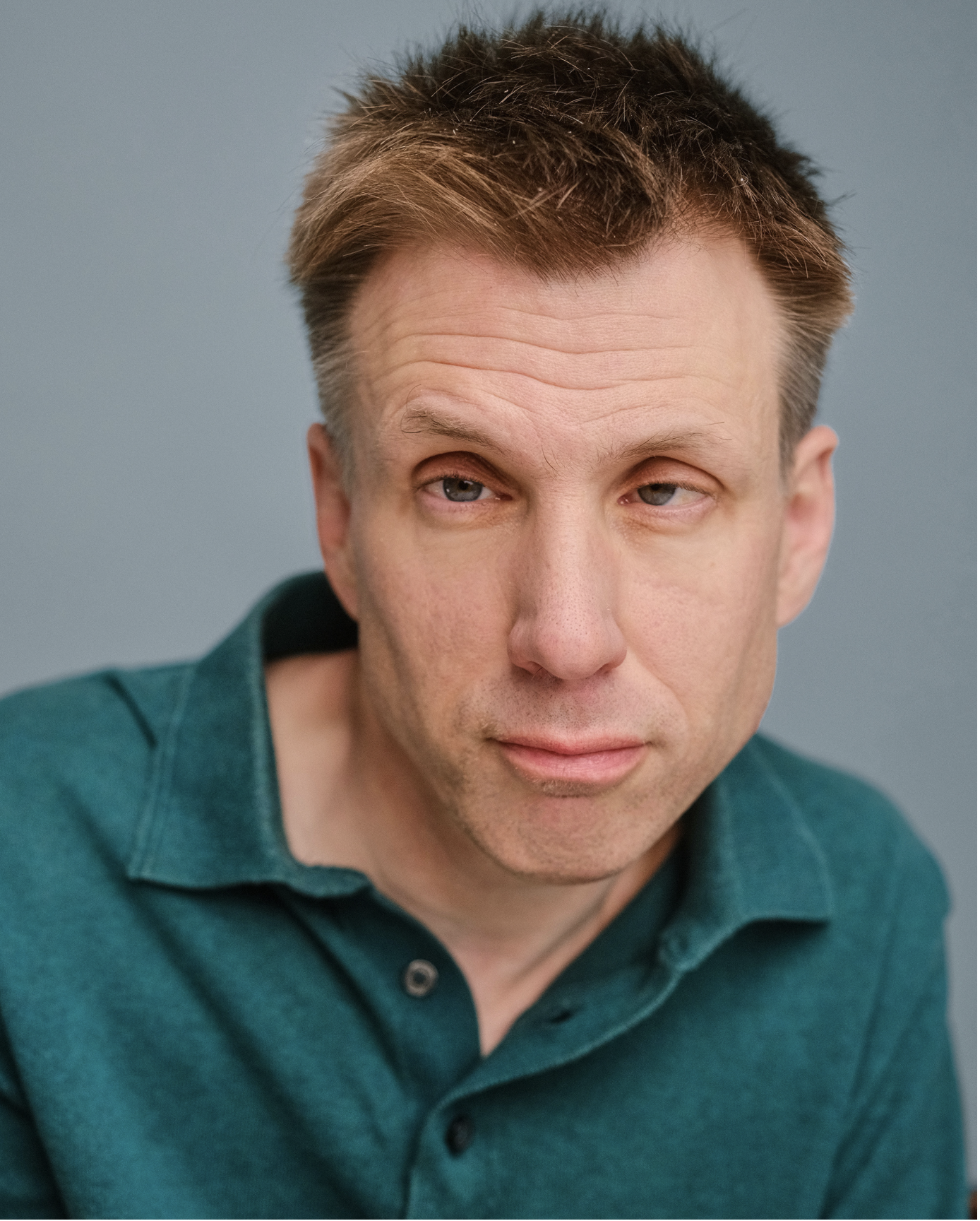 Headshot of actor Tom Housden. Tom is a white man with short brown hair. He is wearing a green shirt and is looking into the camera