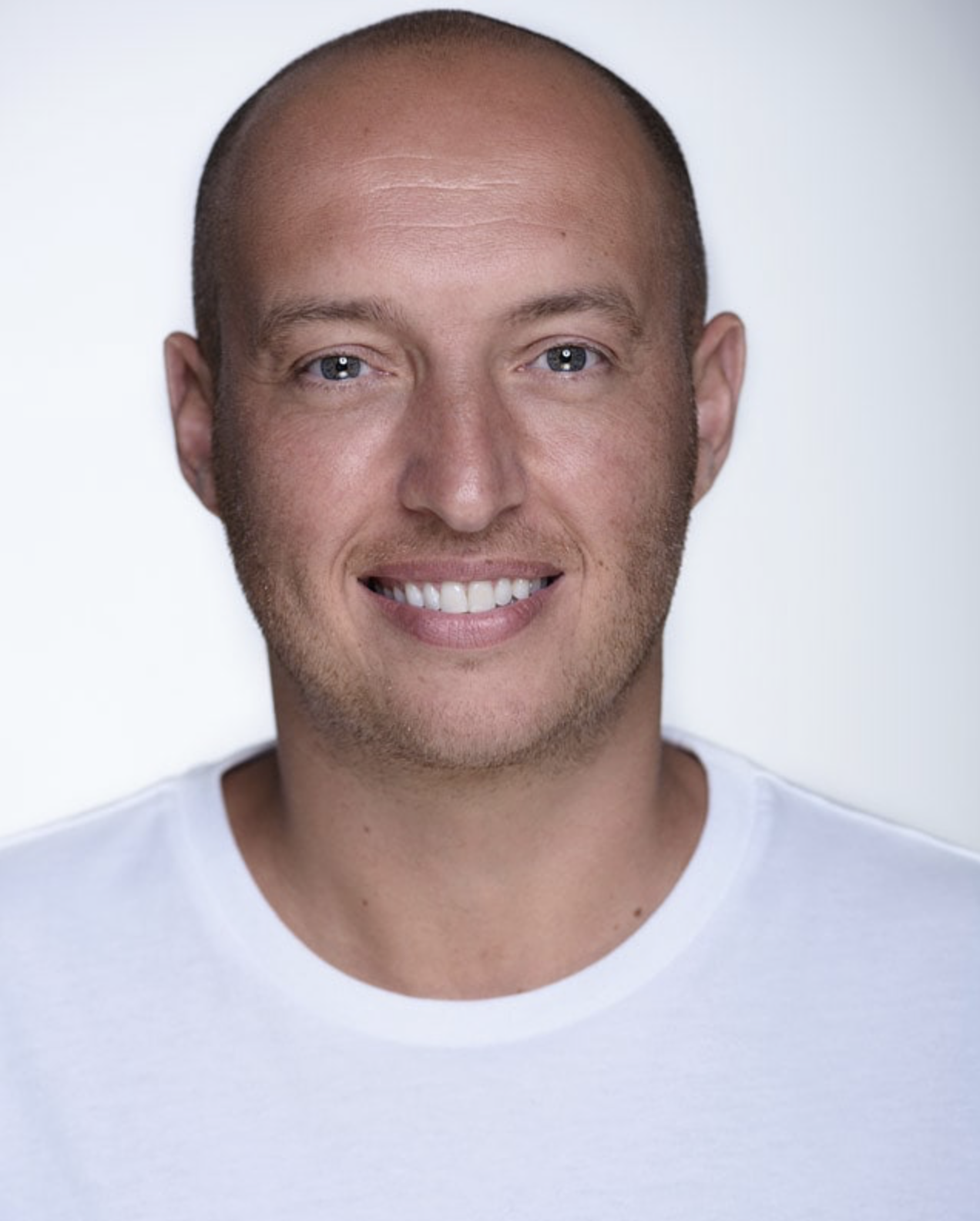 Headshot of actor Tony Bailey. Tony is a white man with a shaved head, green eyes and stubble. He is wearing a white t-shirt. Tony is looking straight at the camera and smiling.