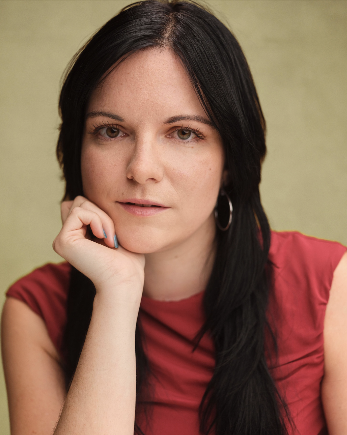 Headshot of actor Abbie Hills. Abbie is a white British woman in her mid-twenties with long, black hair tied up and a fringe. She has brown eyes, long eyelashes, and is looking directly at the camera and smiling. She is wearing a Red top.
