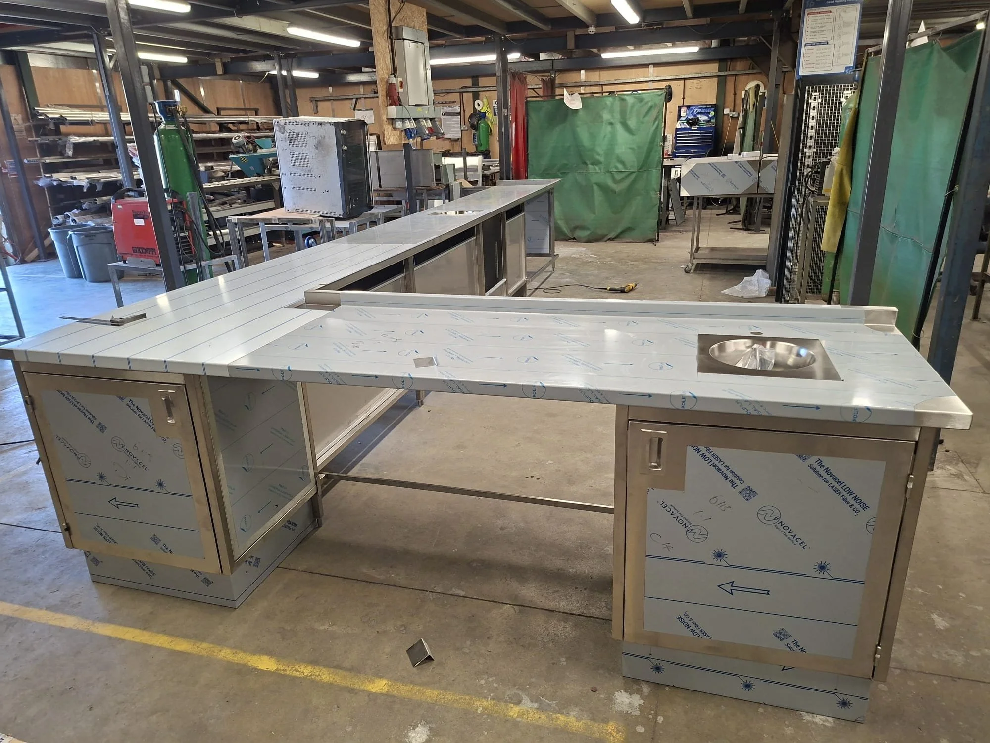 An industrial workshop with metal workbenches, some covered with protective film, and various equipment including a computer and welding tools. There is a green welding curtain in the background, and the space has a high ceiling with exposed beams an
