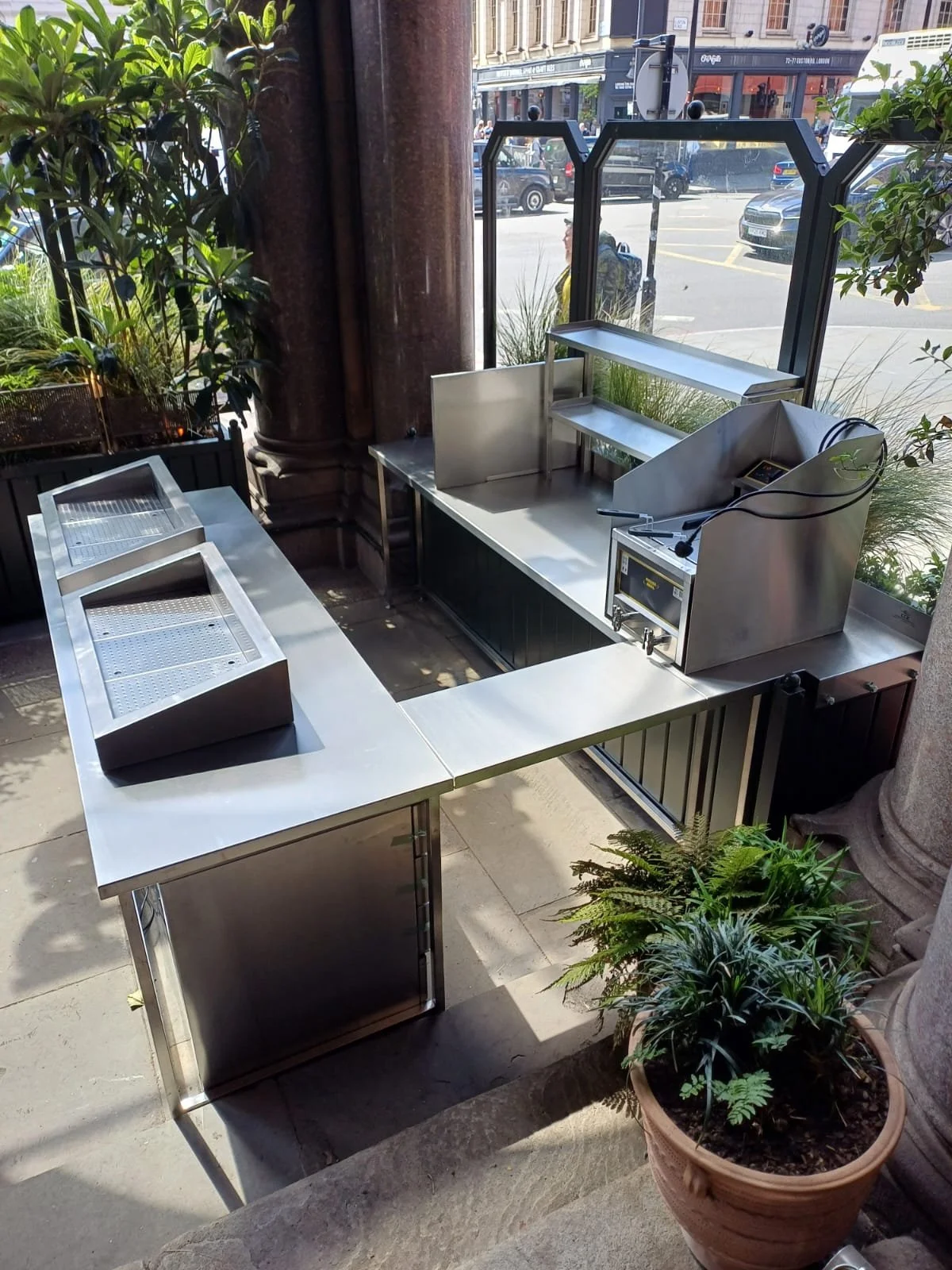 Empty metal food service counter and display trays outside a storefront, with plants and city street in the background.