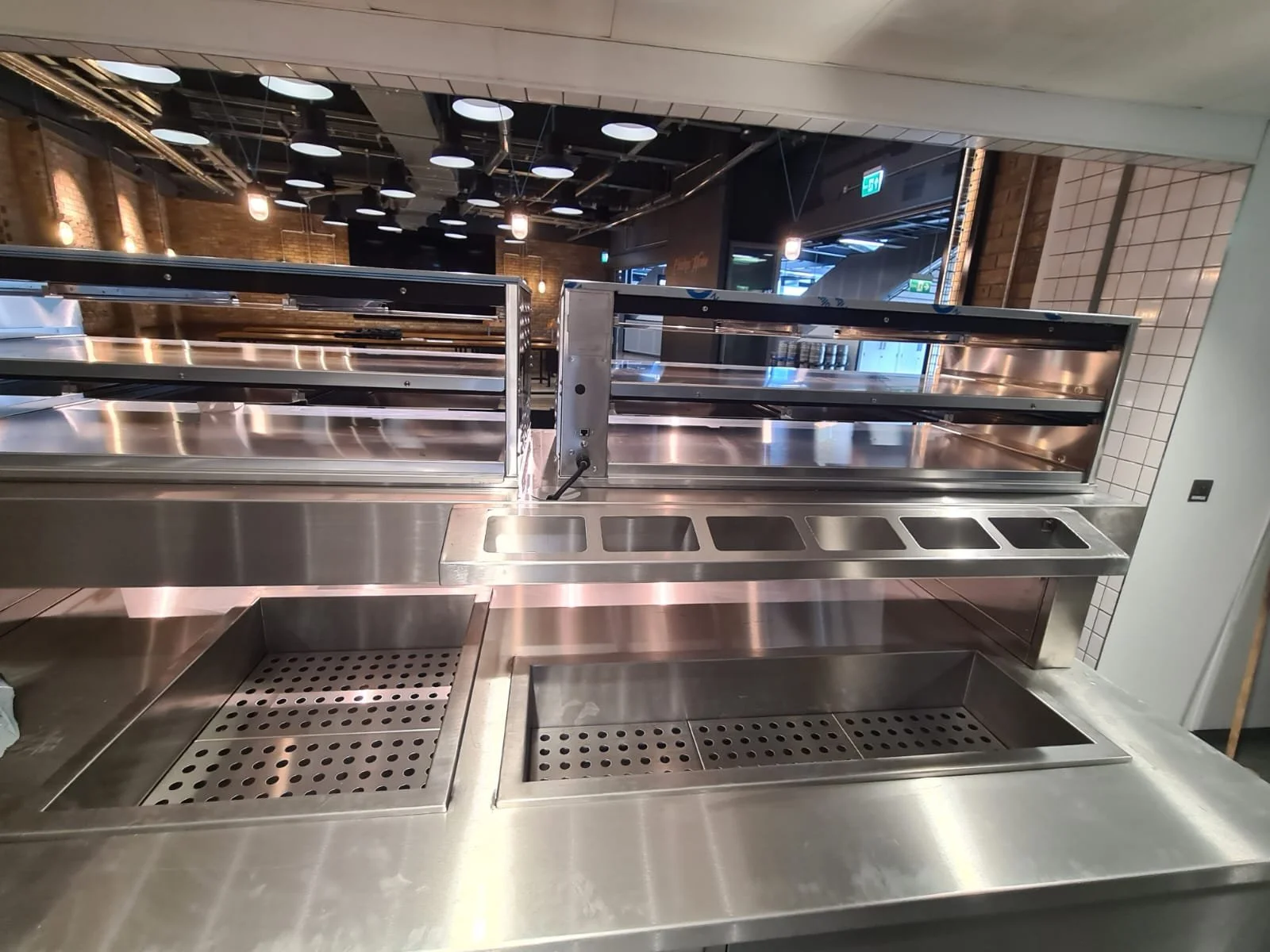 Empty stainless steel food service counter with open refrigerated display case, empty trays, and drainage grates, in a modern restaurant setting.