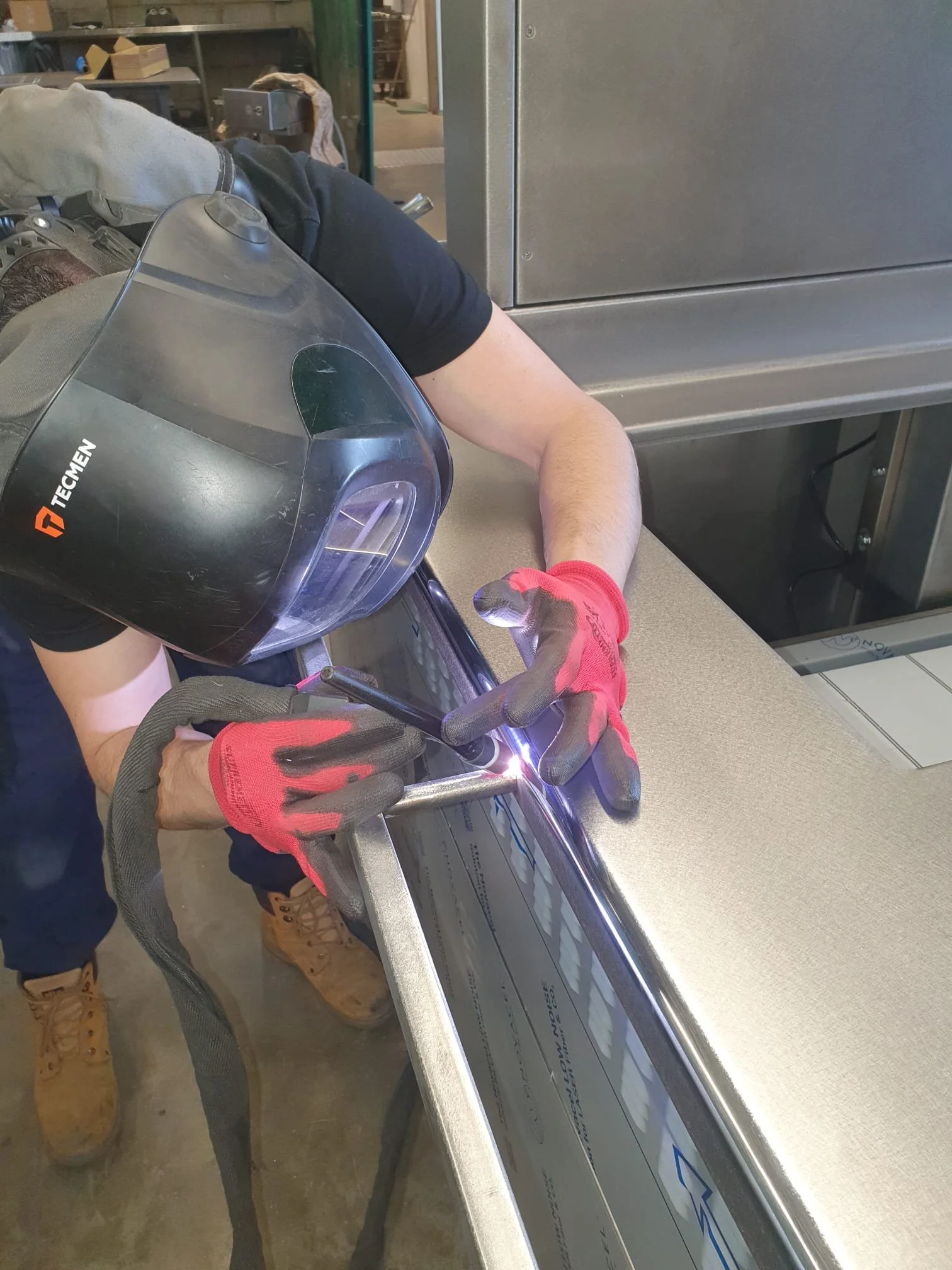 Person welding metal on a workbench inside a workshop, wearing a welding helmet and gloves.