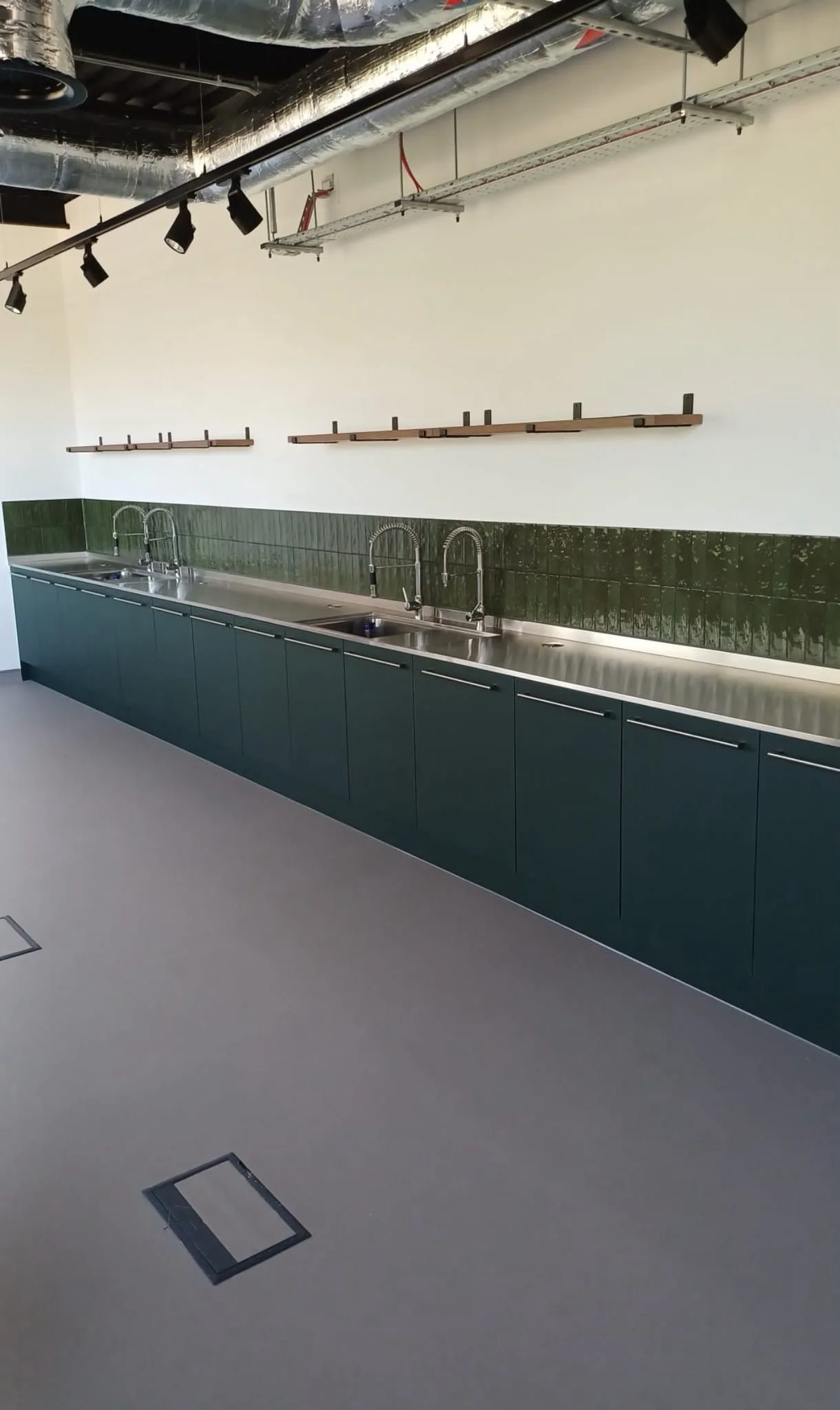 Empty kitchen with green cabinets, stainless steel countertops, three sinks, dark green tile backsplash, and white walls. Ceiling features exposed ductwork and track lighting.